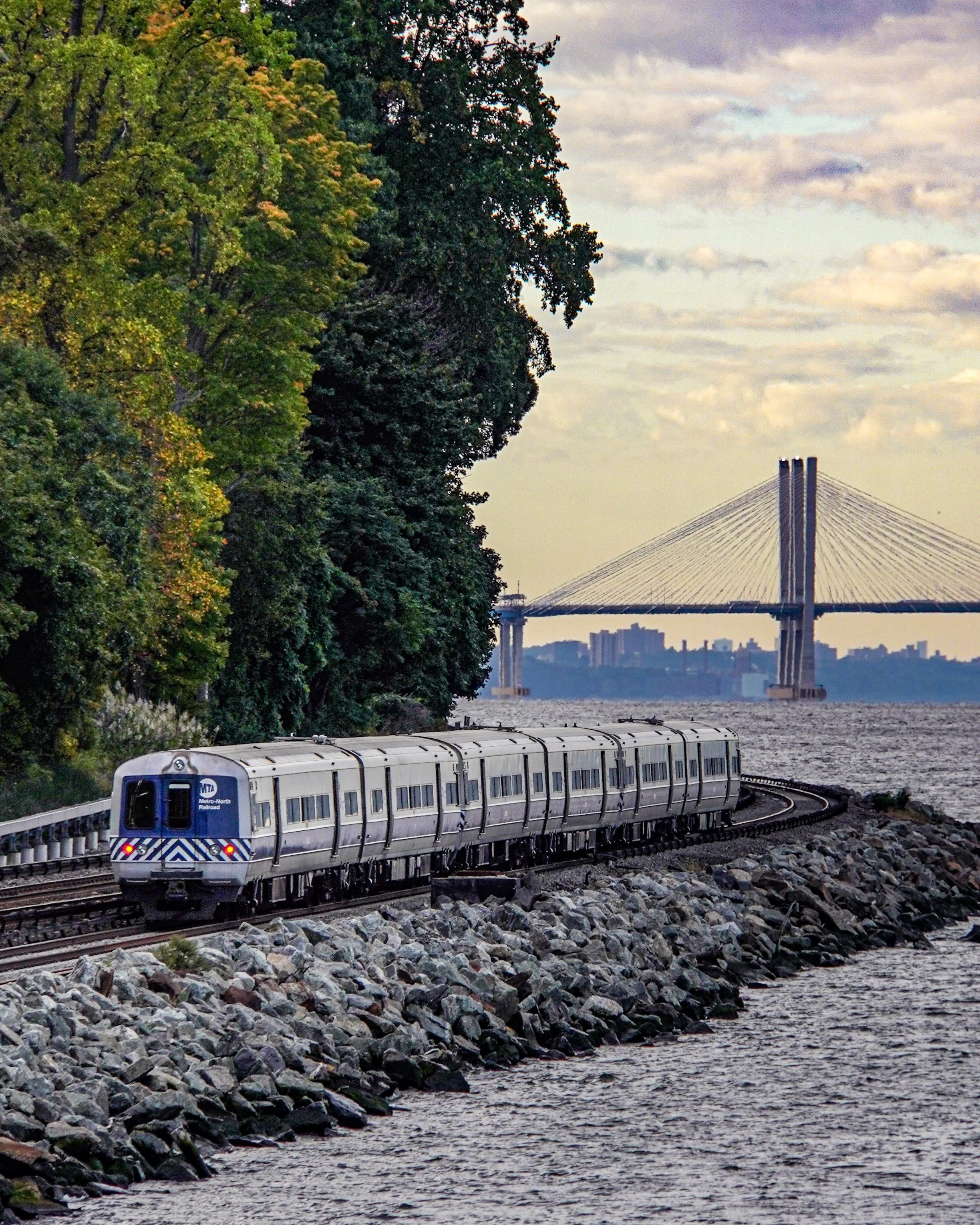 A silver train traveling along a rocky shoreline with trees on the left and water on the right, a bridge with tall cables and cityscape in the background, under a partly cloudy sky.