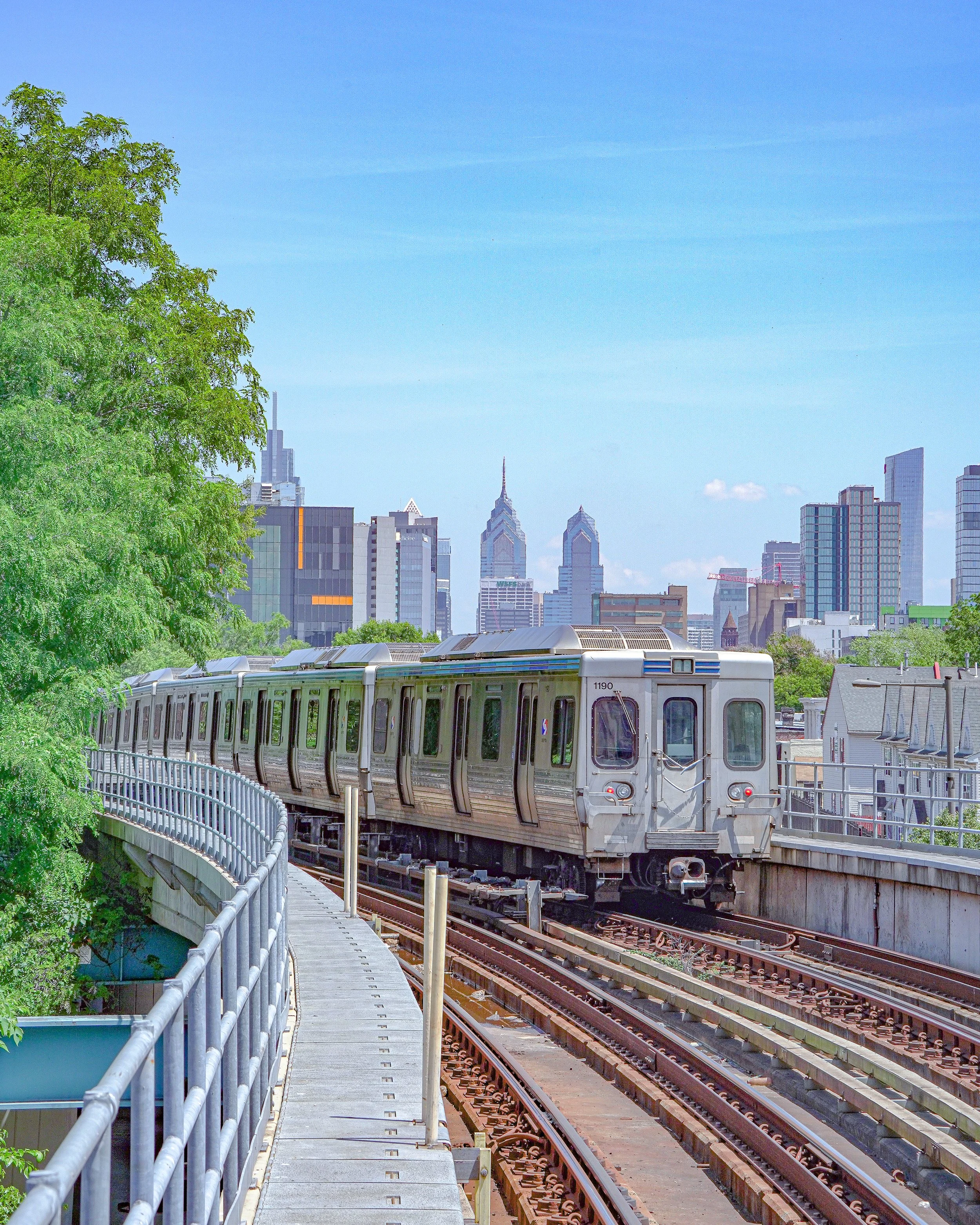 A silver elevated train on tracks with a city skyline in the background, green trees on the left, and a clear blue sky.