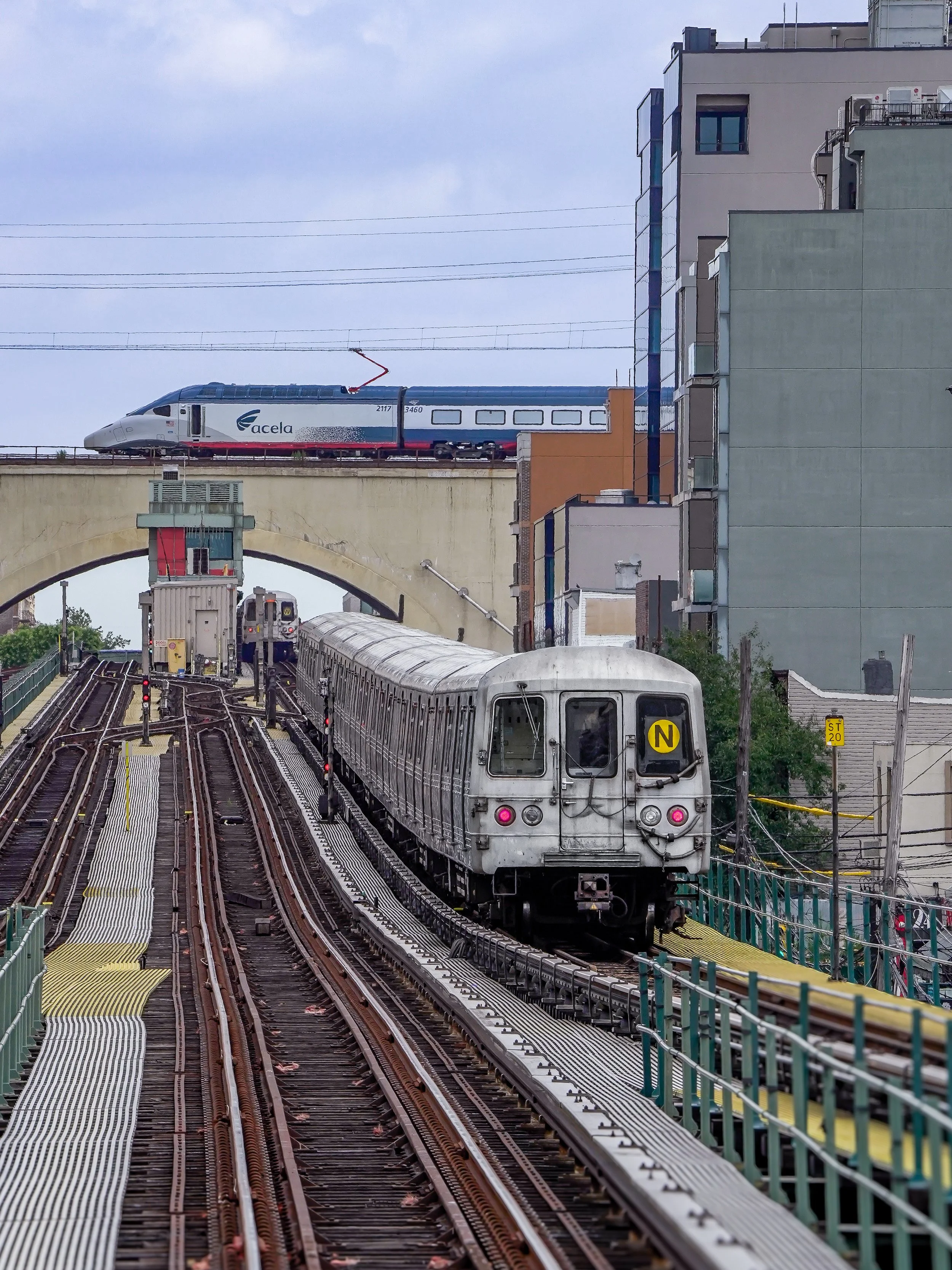 Metro train approaching in an urban environment with a high-speed train on a bridge above.