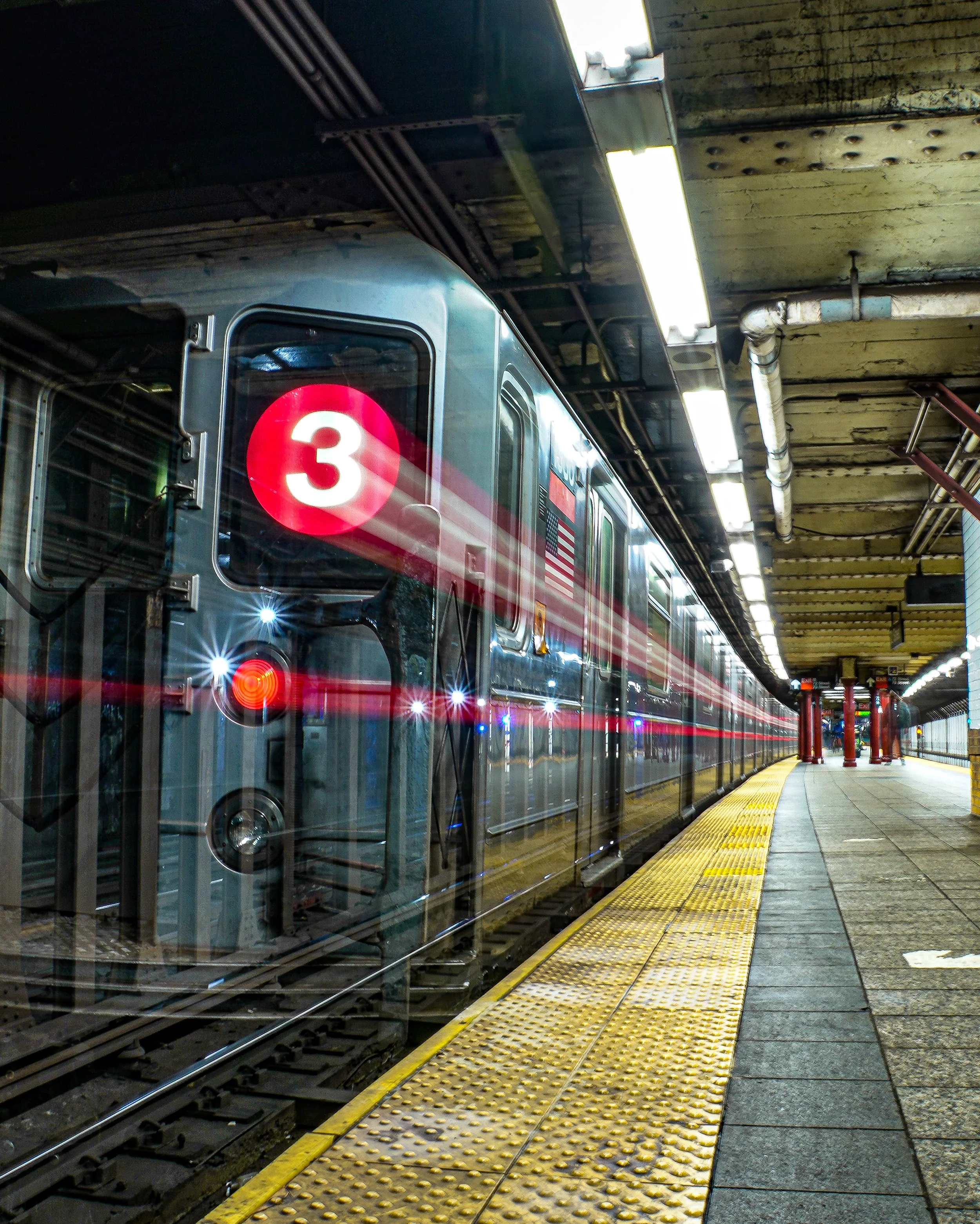 An underground train station platform with a silver subway train departing the station, displaying '3' on the front. The platform has a yellow tactile paving strip along the edge and a row of fluorescent lights on the ceiling.
