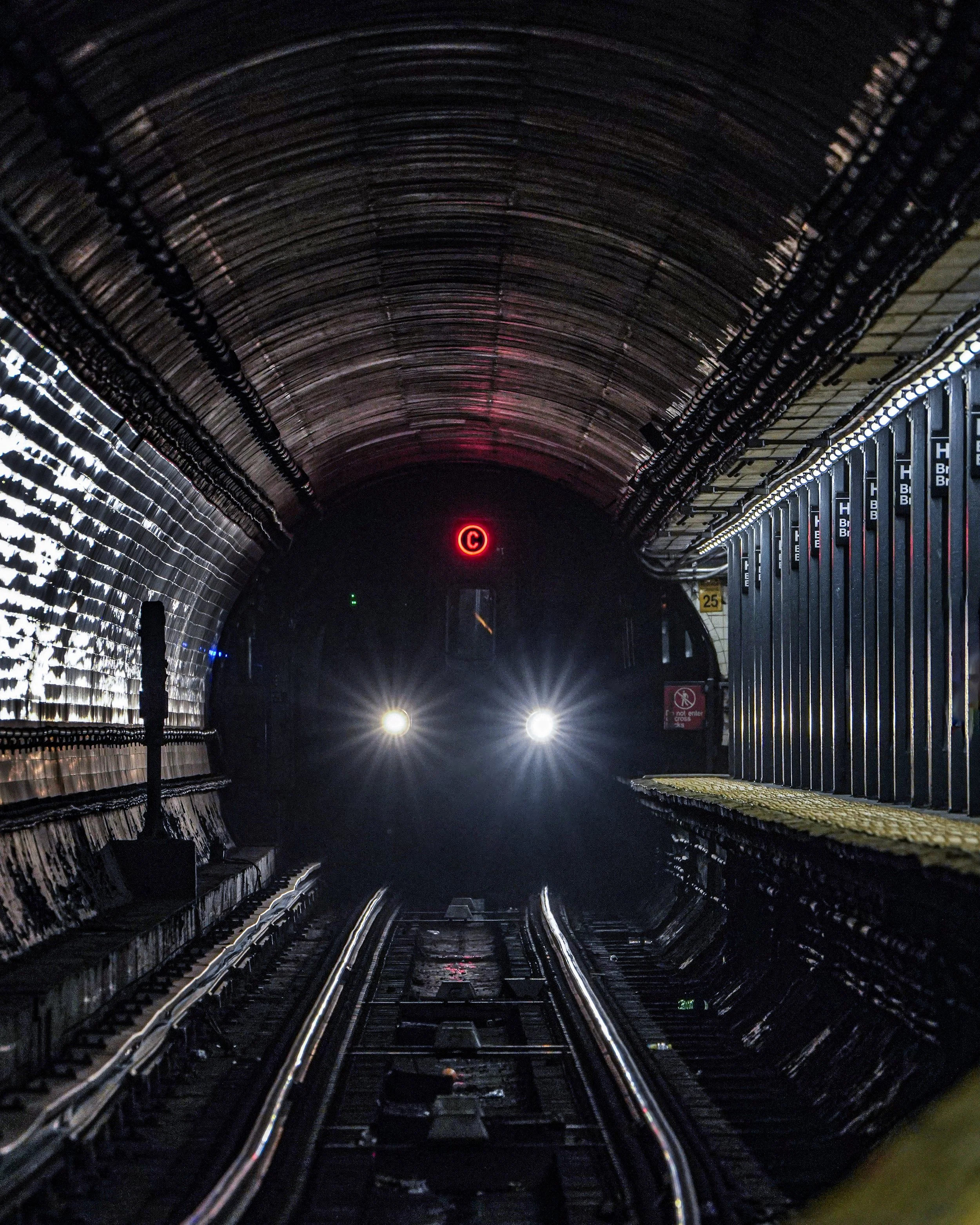 Inside a dark subway tunnel with an approaching train having bright headlights and a red 'C' signal above.