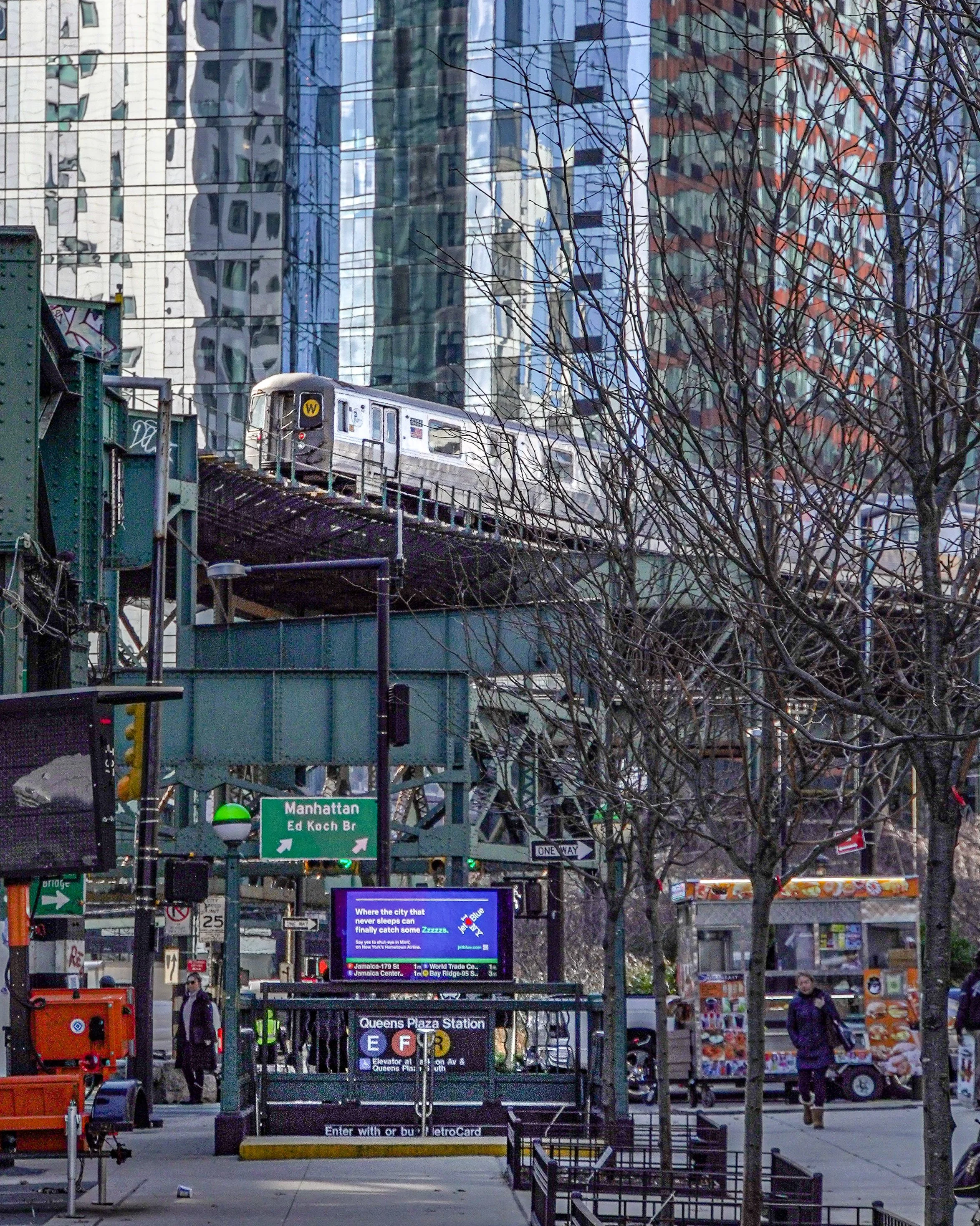 An elevated train passing over a train track in a city, with modern glass skyscrapers in the background, trees in the foreground, and a subway entrance with signs for Manhattan and Queens Plaza Station below.