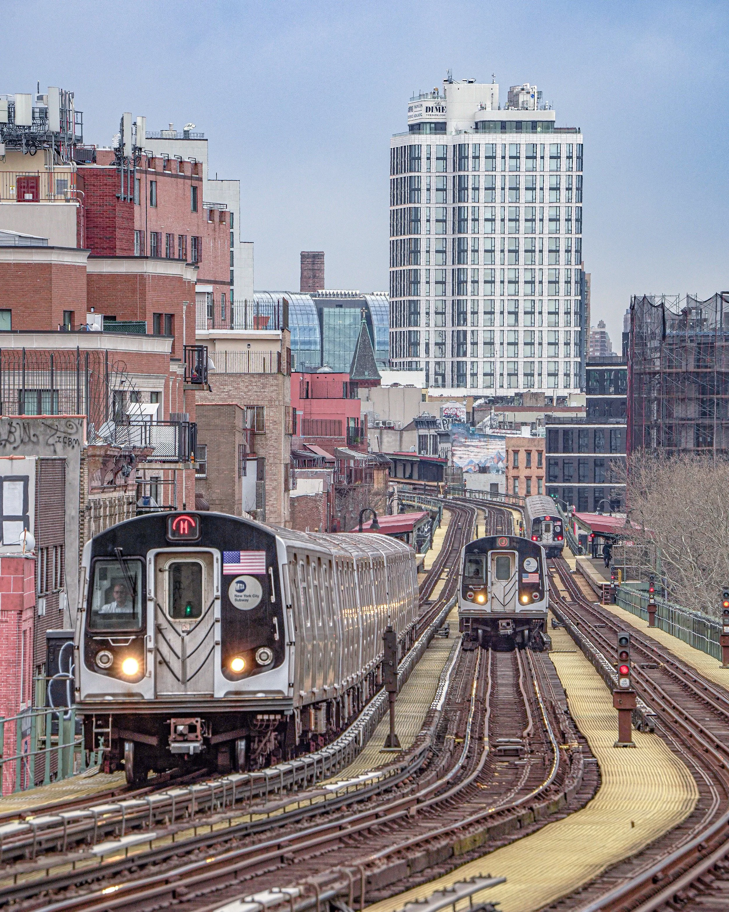 View of New York City elevated subway trains on tracks with urban buildings and skyscrapers in the background.