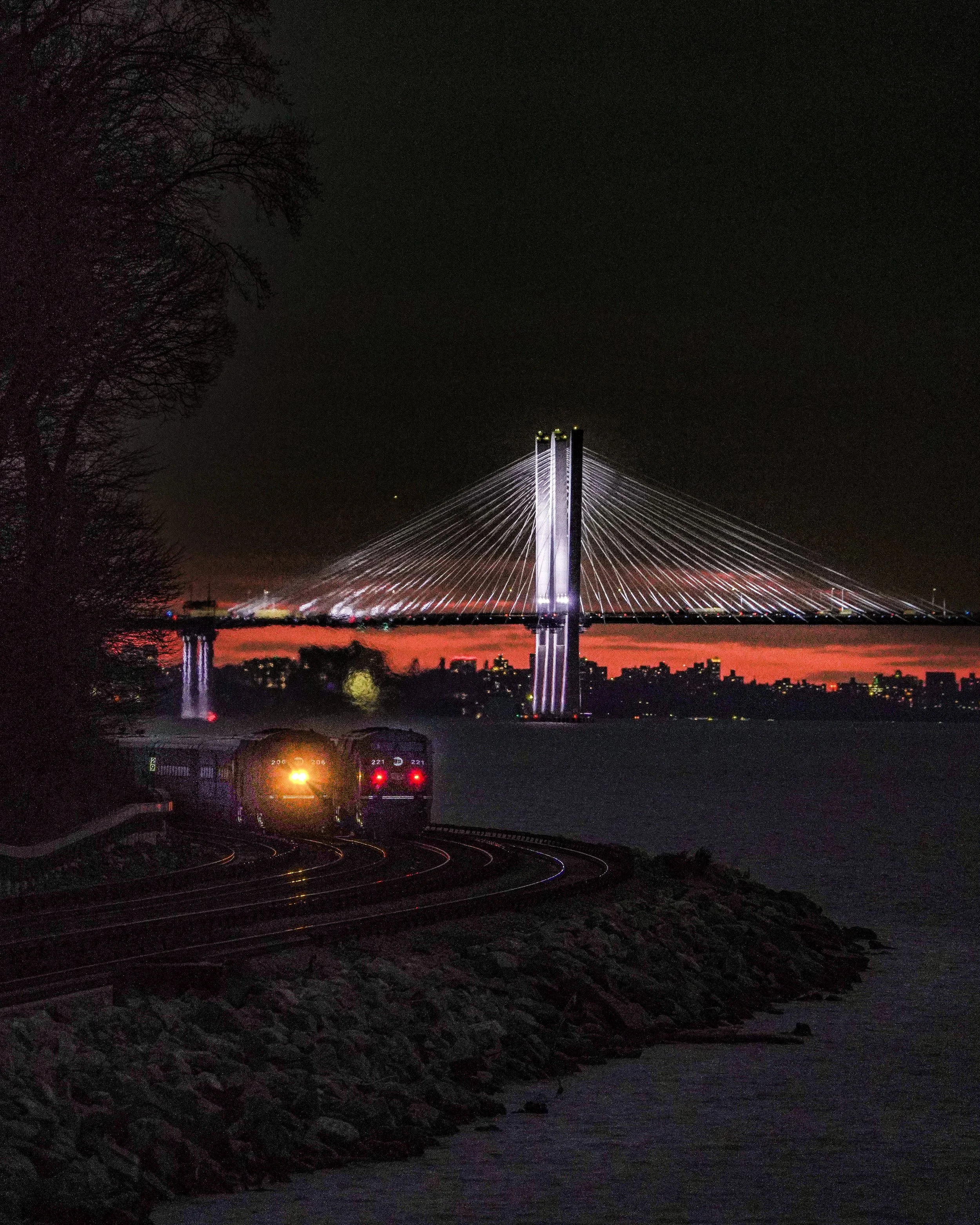 Nighttime view of a illuminated cable-stayed bridge over a body of water, with a train on curved tracks in the foreground and a city skyline in the distance.