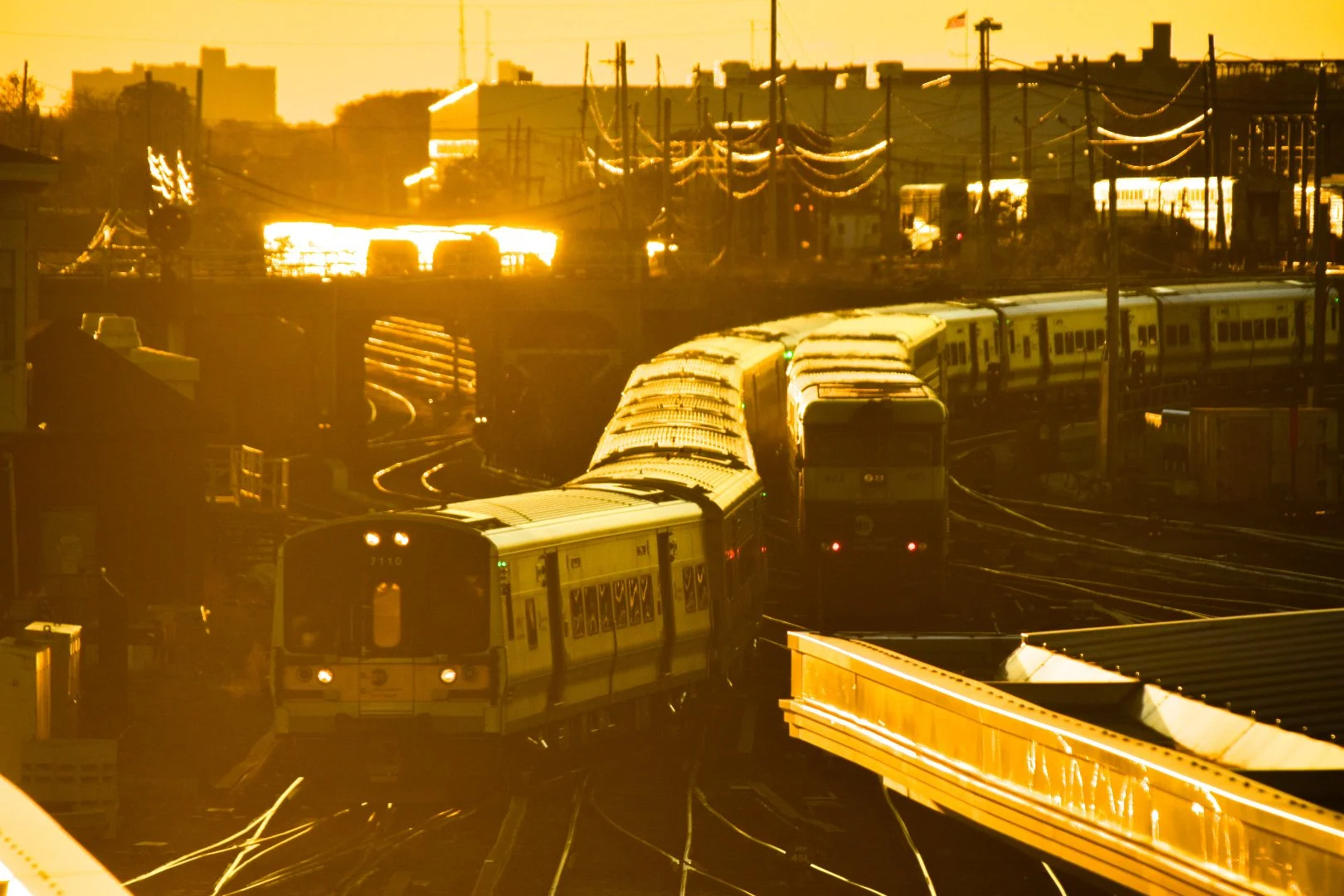 A city train on the tracks during sunset or sunrise with a golden glow, power lines overhead, and buildings in the background.