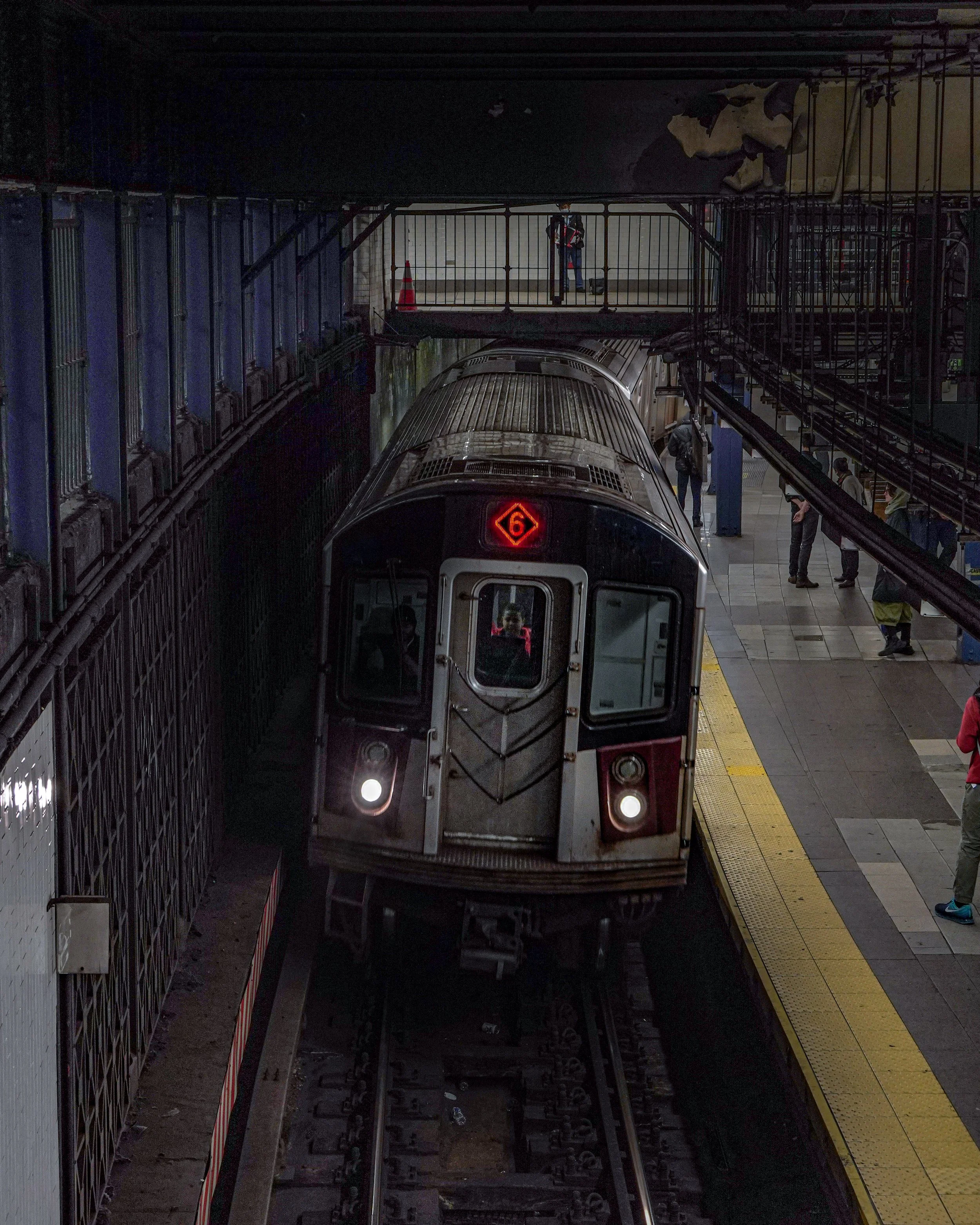 An underground train station with a train on the track and passengers on the platform, with a pedestrian walking on a bridge above.