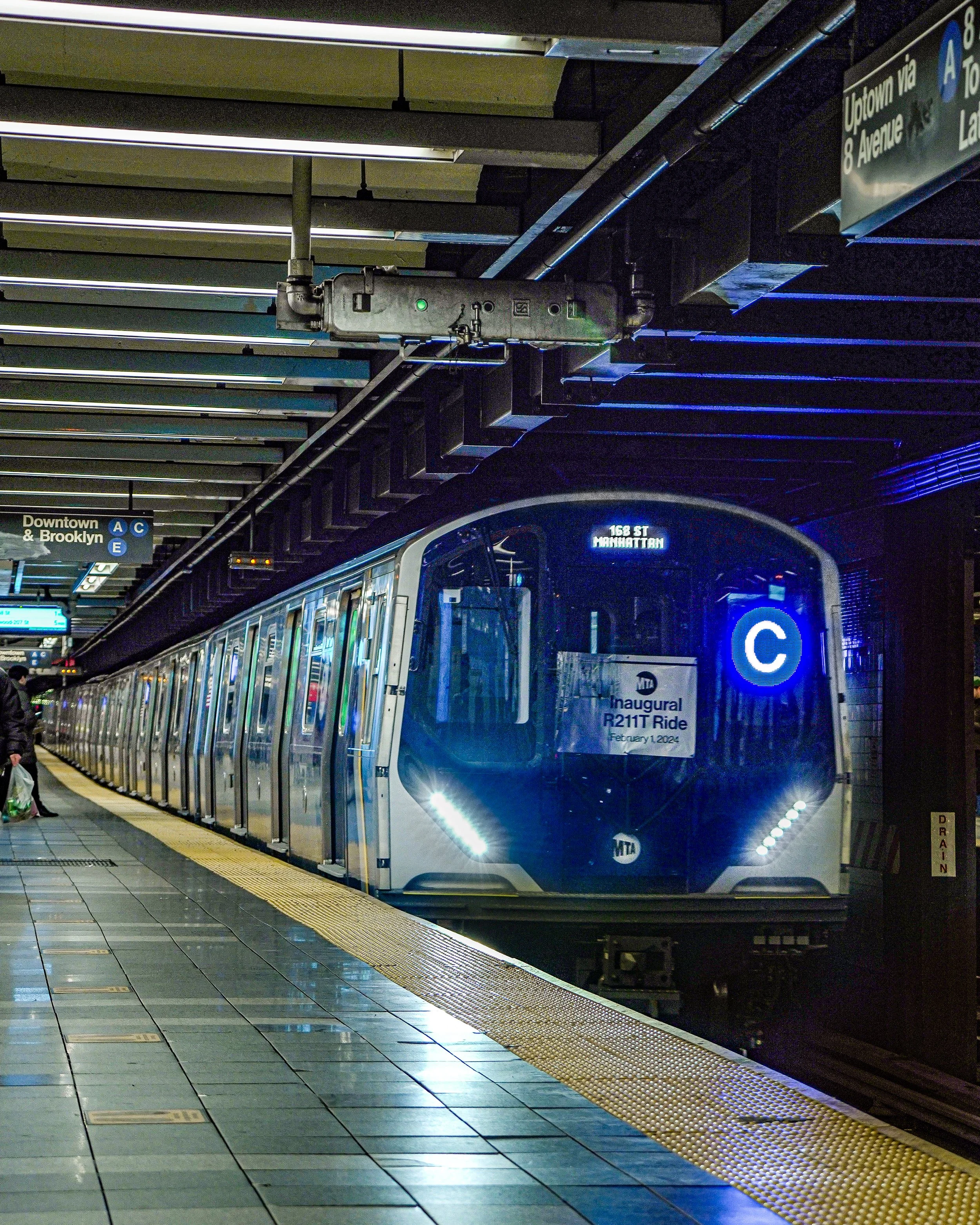 A subway train at a station with a digital blue 'C' sign on the front, indicating the train's line, with illuminated signs and passengers on the platform.