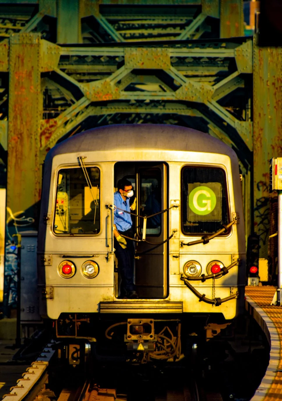 A train operator wearing a mask and gloves stands inside the front of a subway train marked with a green 'G' symbol, approaching or going under a metal bridge structure.