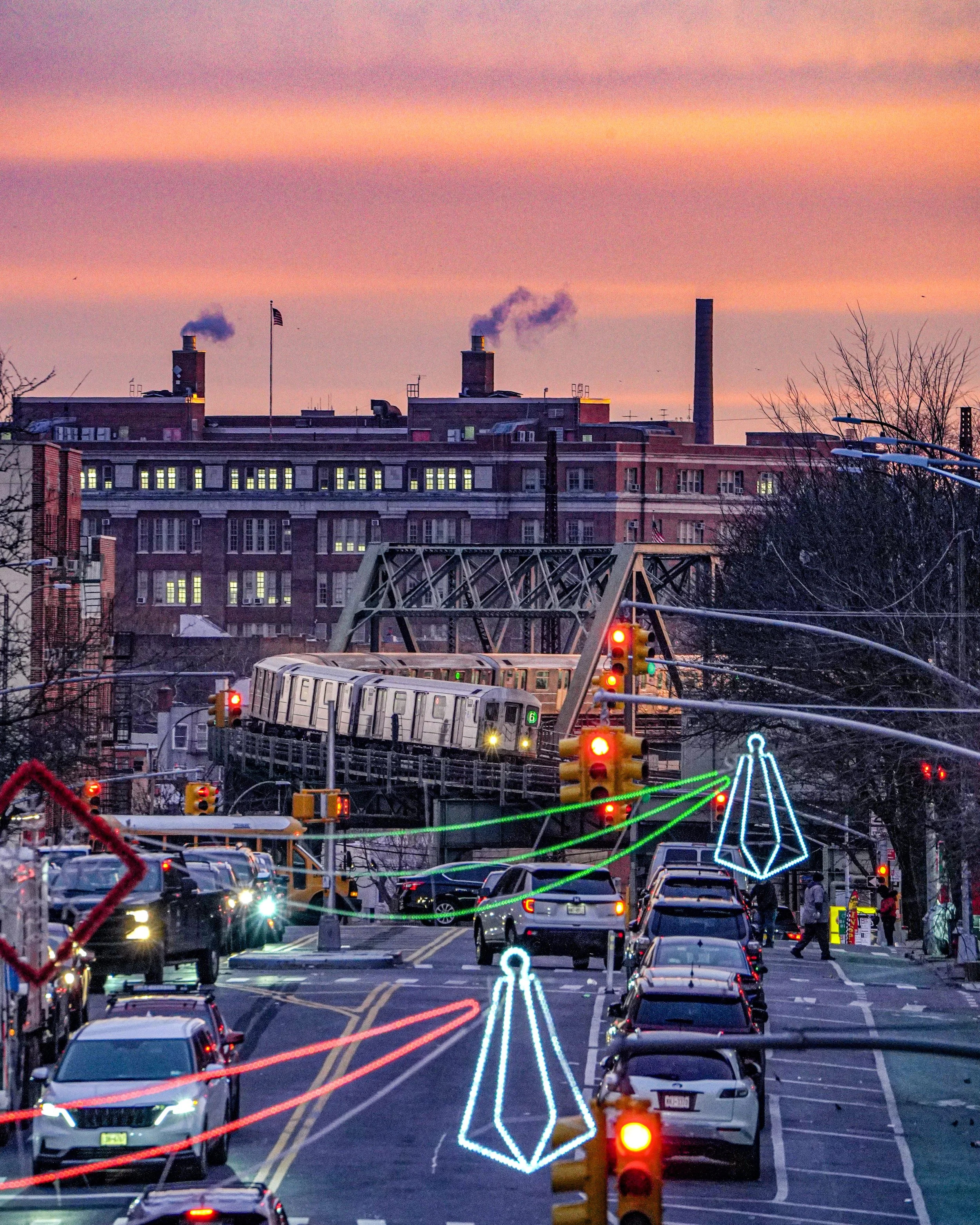 City street with parked cars, traffic lights, holiday decorations, a train in the background, and a brick building with smoke coming out of chimneys at sunset.