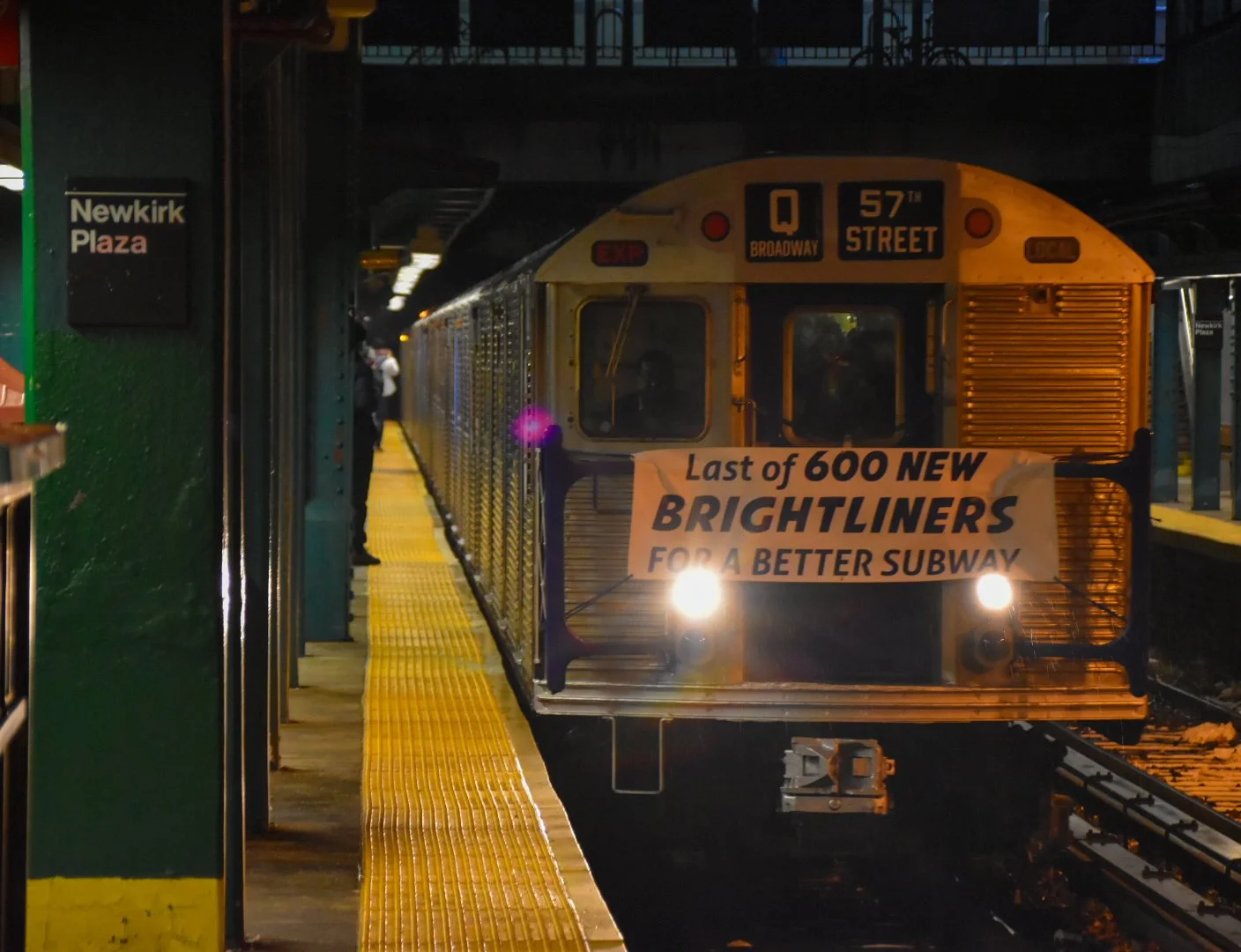 A New York City subway train stopped at the platform with a sign on the front that reads, "Last of 600 new Brightliners for a better subway." The train displays the destination as 57th Street on the Q Broadway line. The platform is illuminated with y