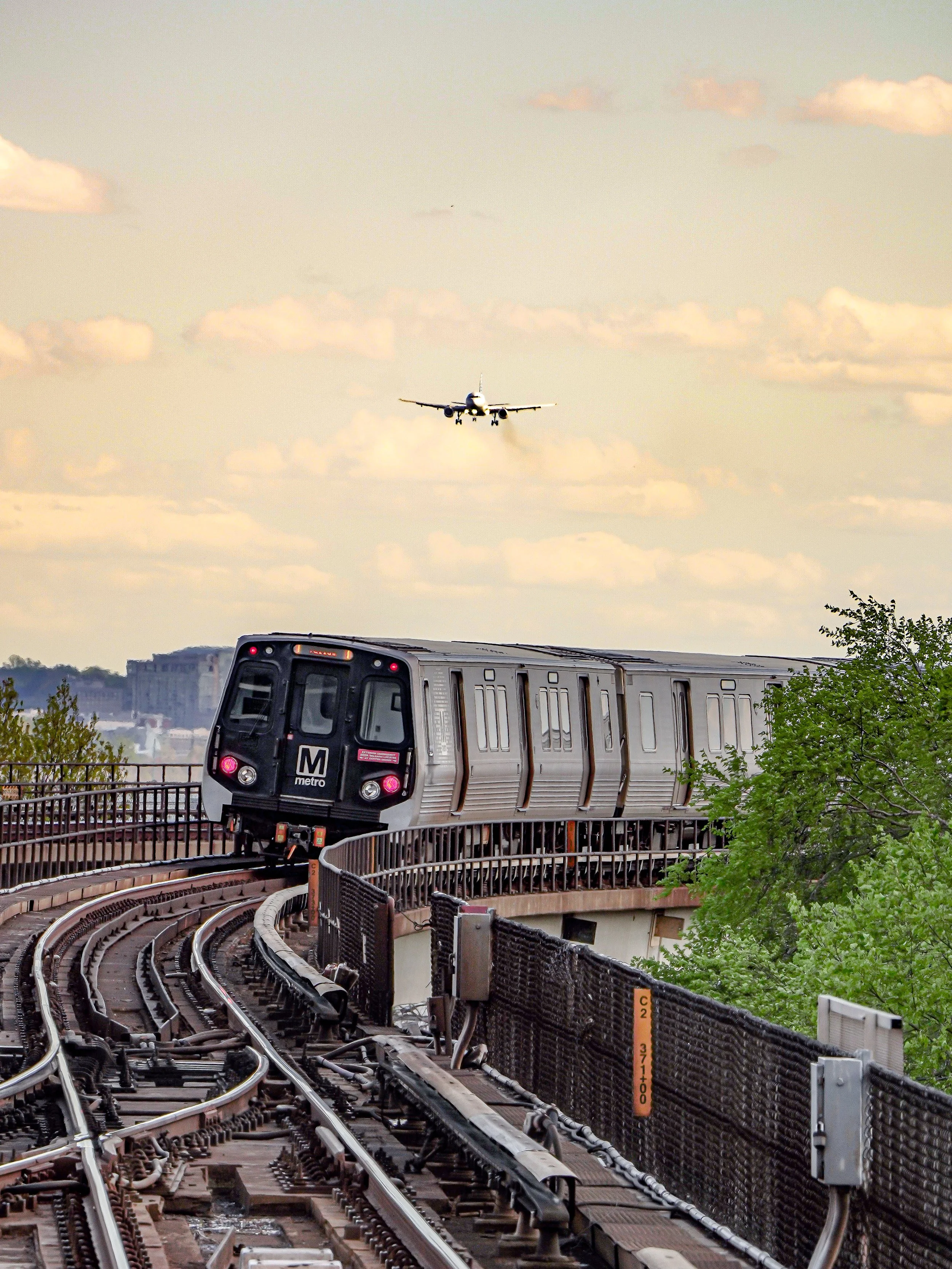 A metro train on an elevated track with an airplane flying in the sky above.