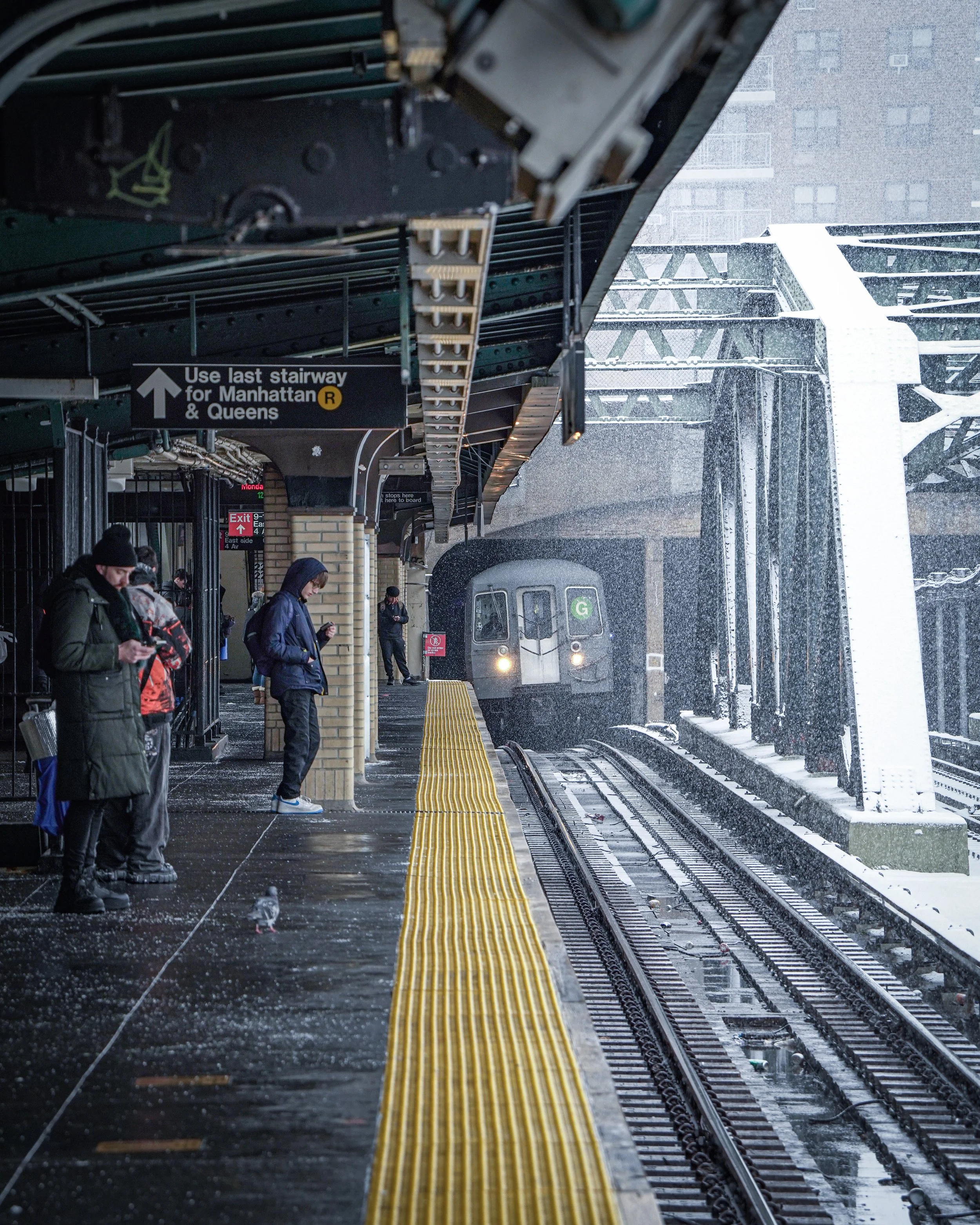 People waiting at a snowy subway station platform as a train arrives. The scene includes commuters dressed in winter clothing and a train approaching in the distance.