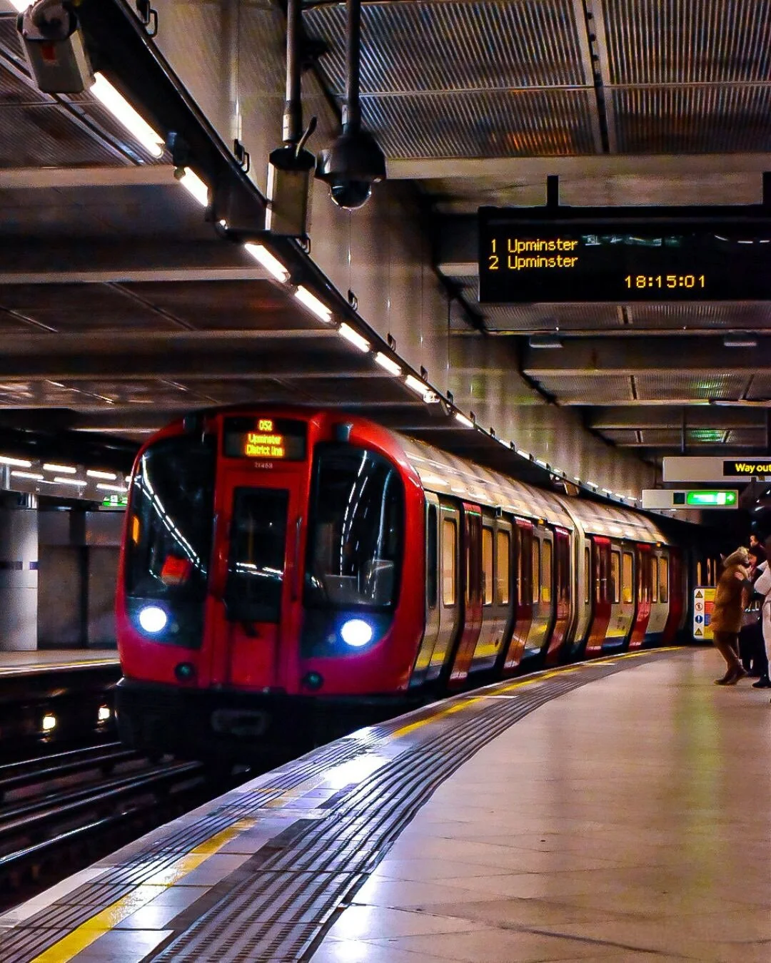 A red London Underground train arriving at the station platform, with a digital display showing its destination and departure time, and passengers waiting nearby in an indoor subway station.