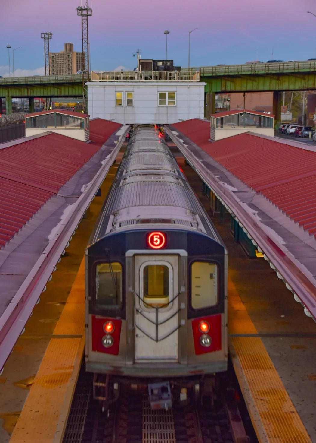 A train on an outdoor platform with a red stop sign displaying the number 5, under a pink and blue sky, with elevated green rail tracks and tall buildings in the background.