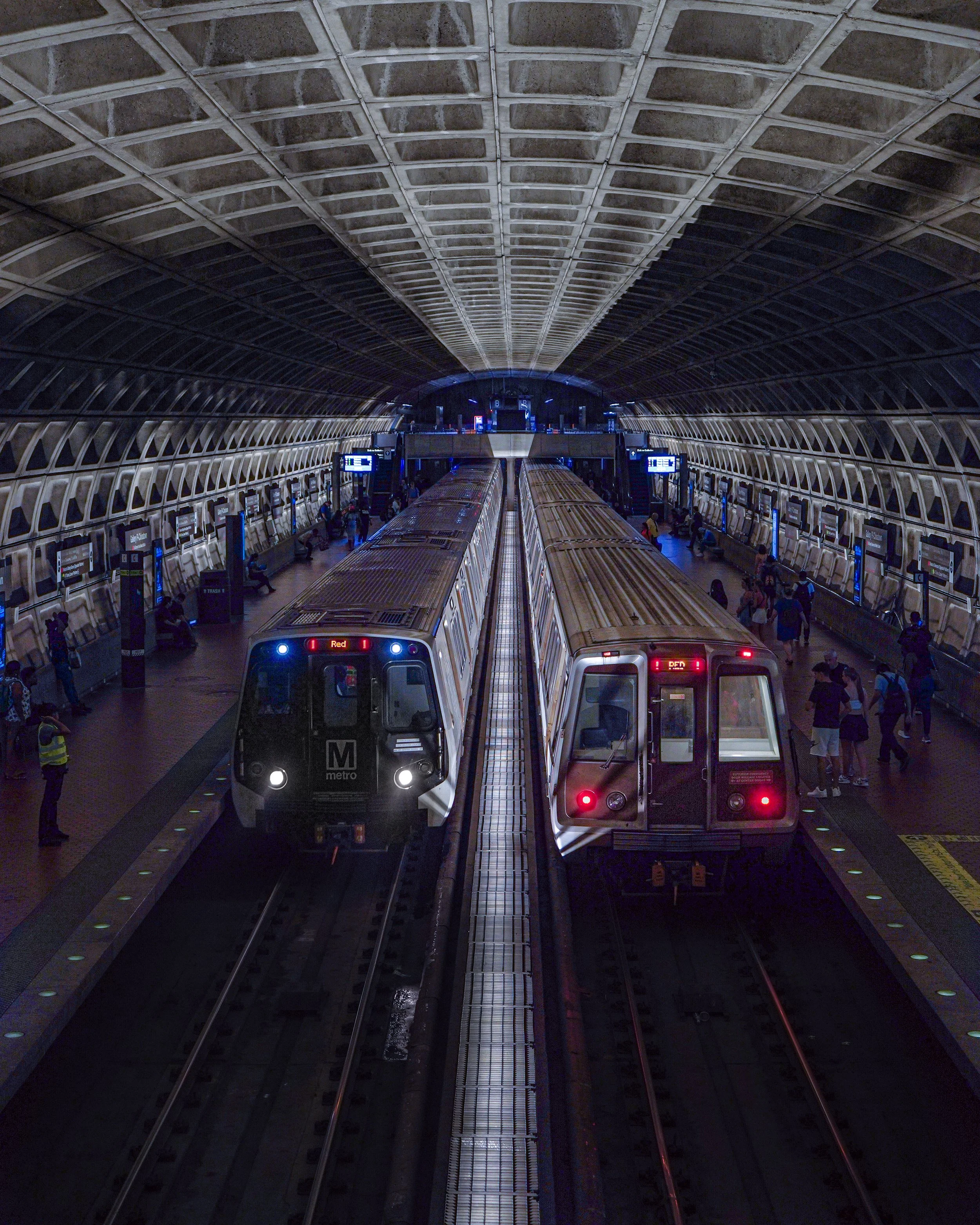 Inside a large metro station with two trains on parallel tracks and passengers waiting on the platform.