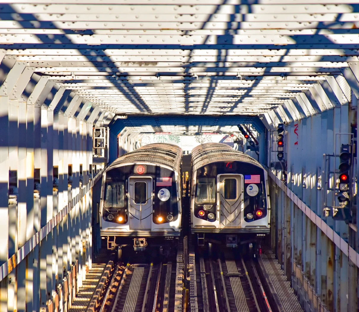 Two subway trains on elevated tracks passing through a metal tunnel with shadows cast by the structure.