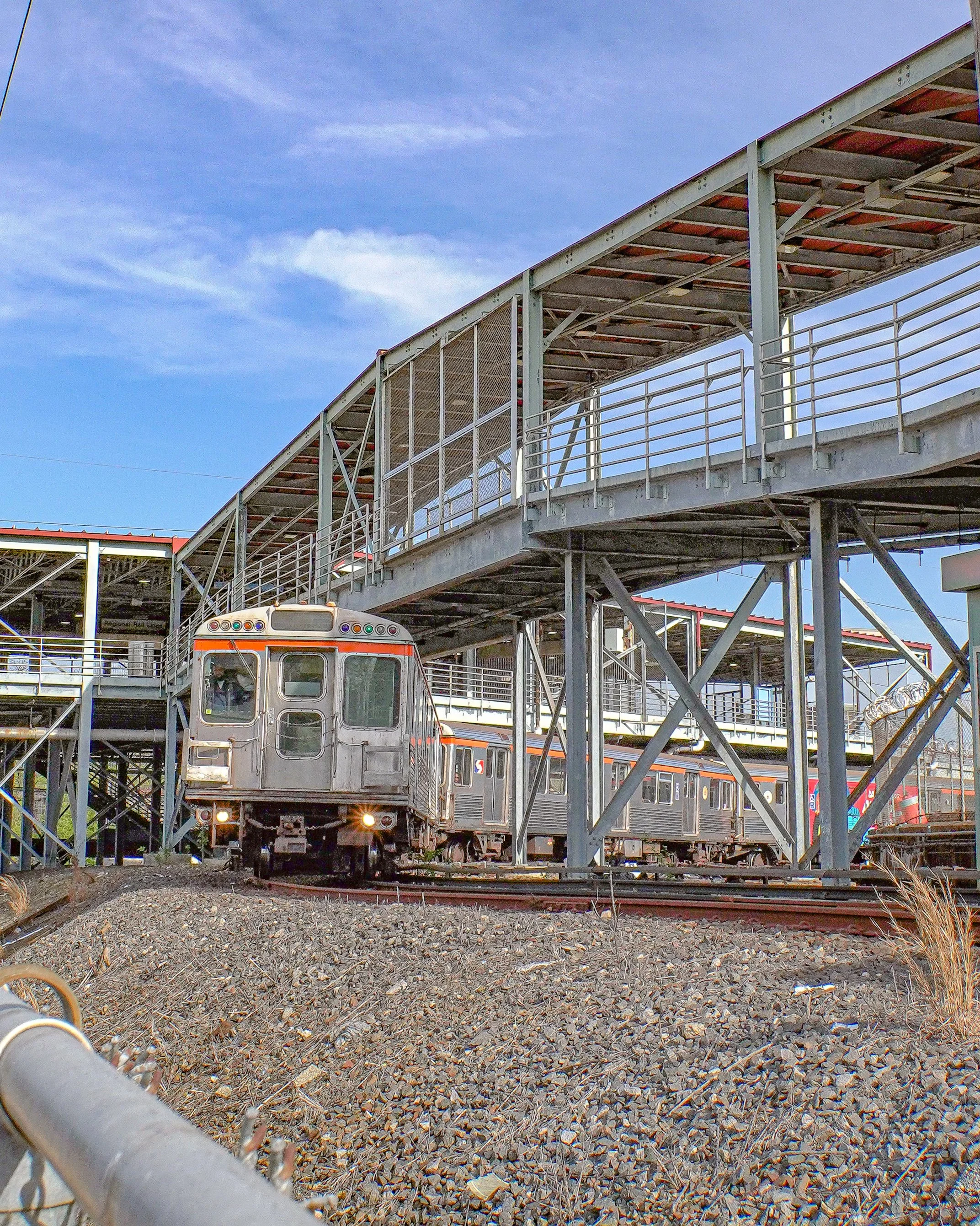 A silver train traveling on tracks beneath a metal pedestrian overpass on a clear day.