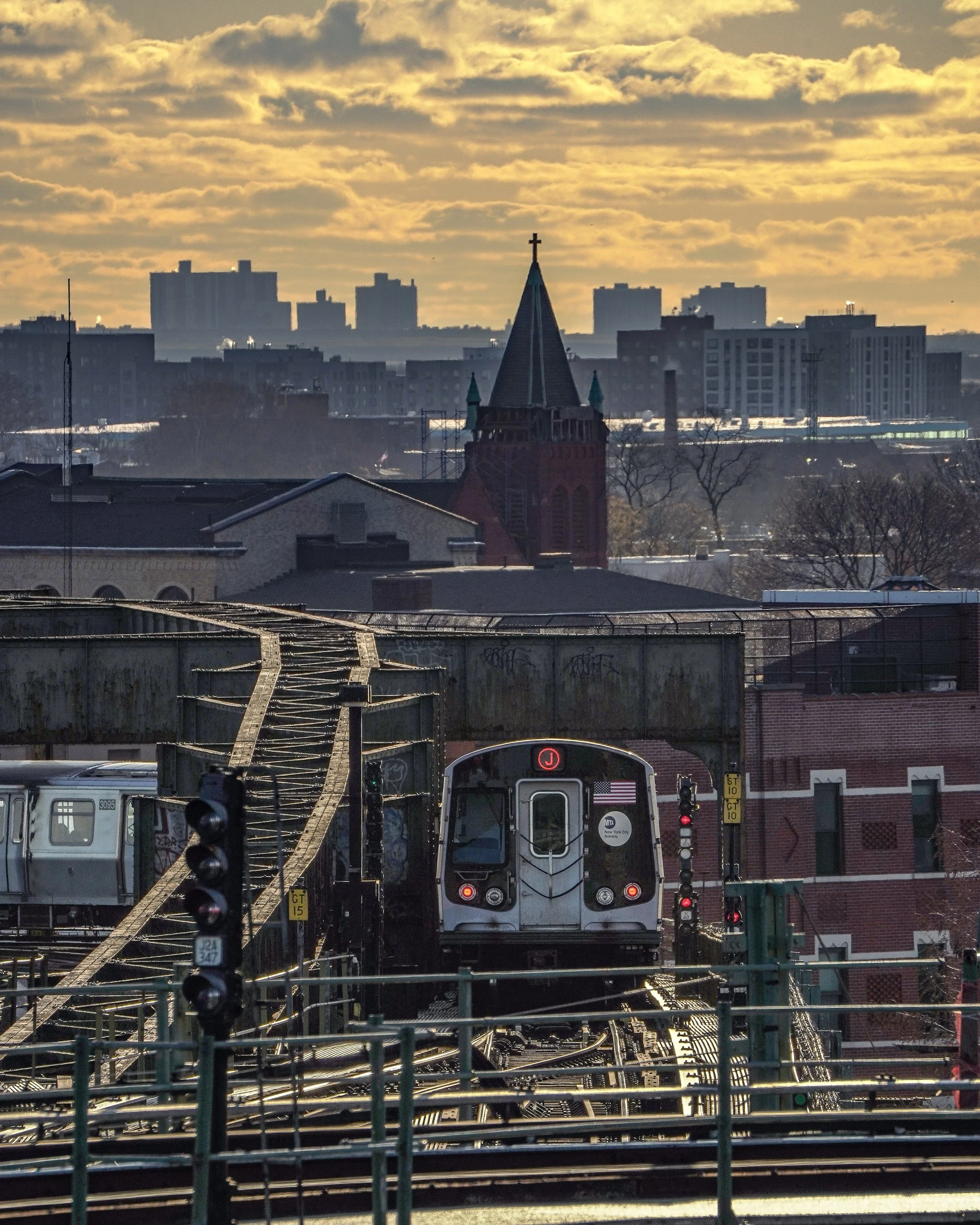 A train on elevated tracks with city buildings and a church with a tall steeple in the background during sunset.