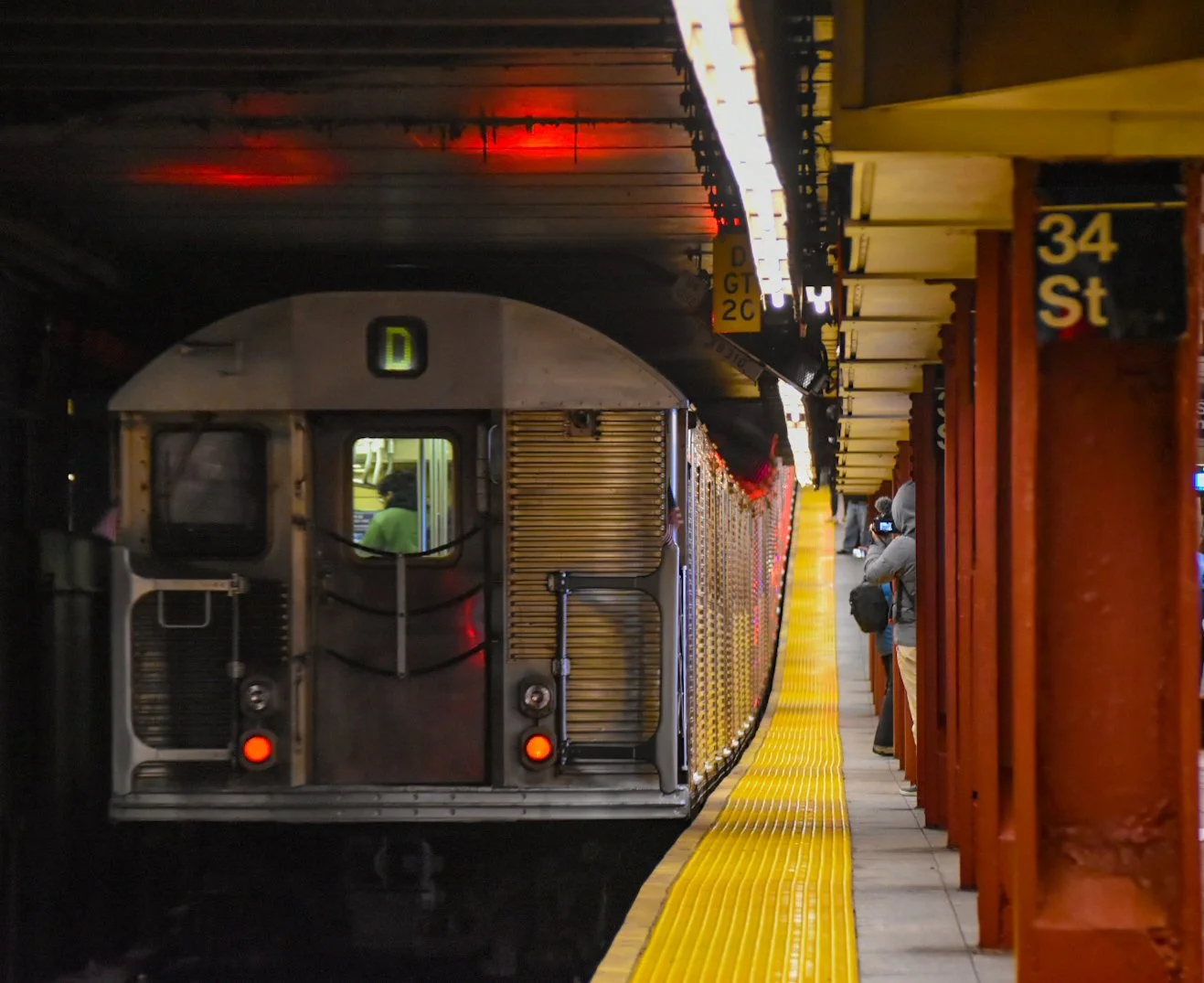 San Francisco Bay Area BART train stopped at underground station platform with passengers waiting to board, illuminated yellow tactile paving along edge of platform, orange support columns, and trajectory view of the train.