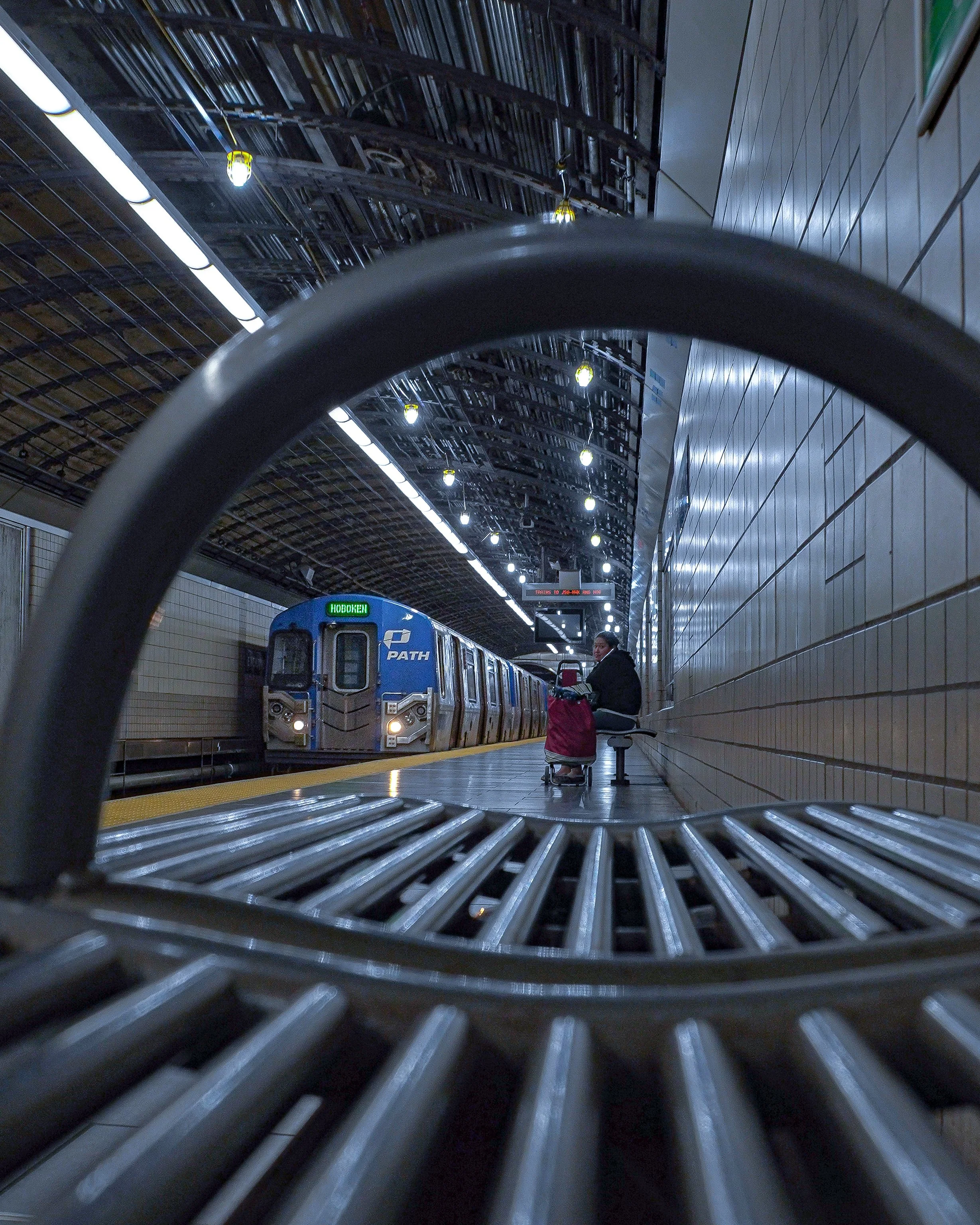 View of a subway station platform with a person sitting on a bench and a train approaching, seen through a metal grate with a low angle shot.