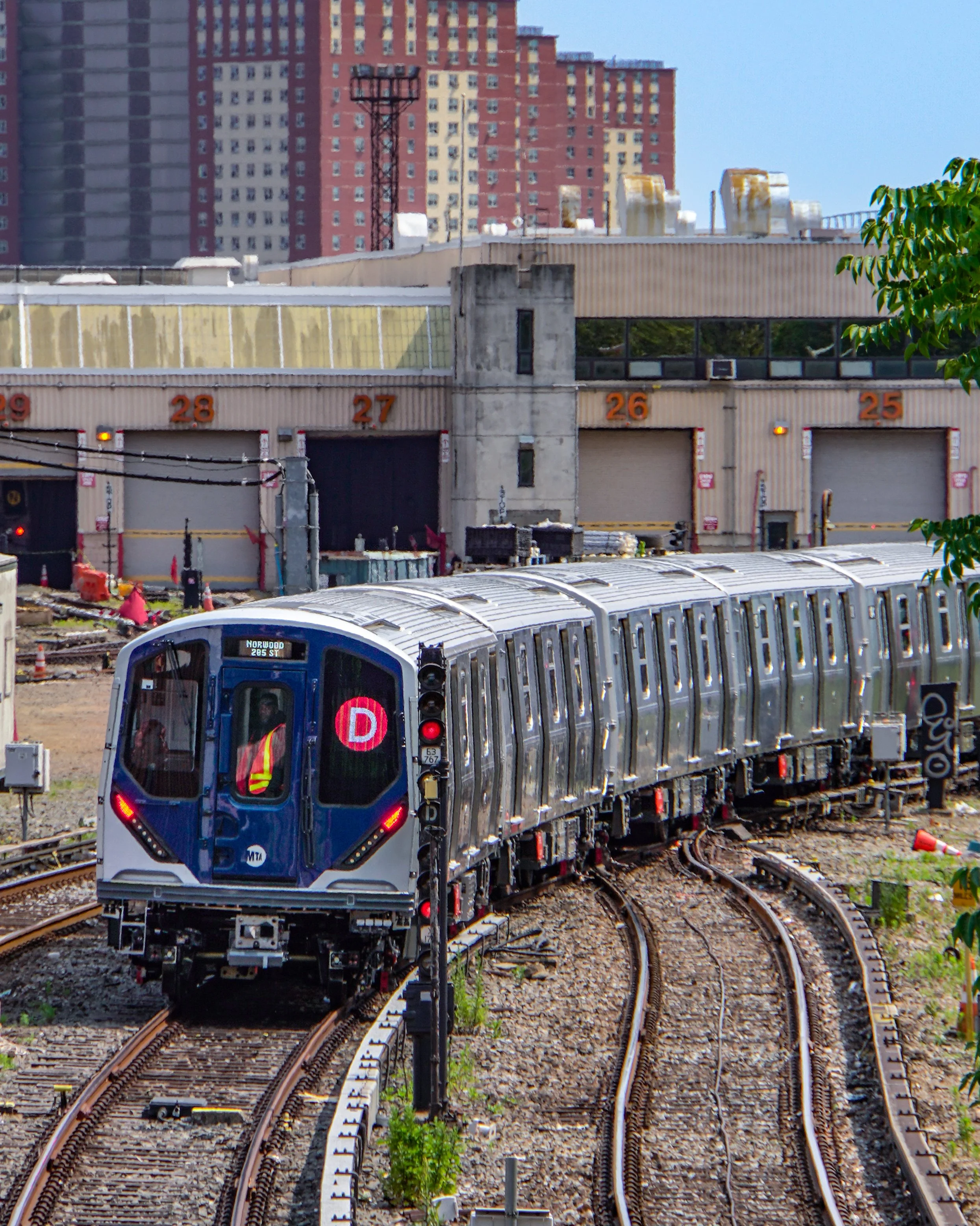 A modern subway train with a digital sign indicating the D train heading to the Coney Island yard, with an urban background of multi-story buildings and a construction site.
