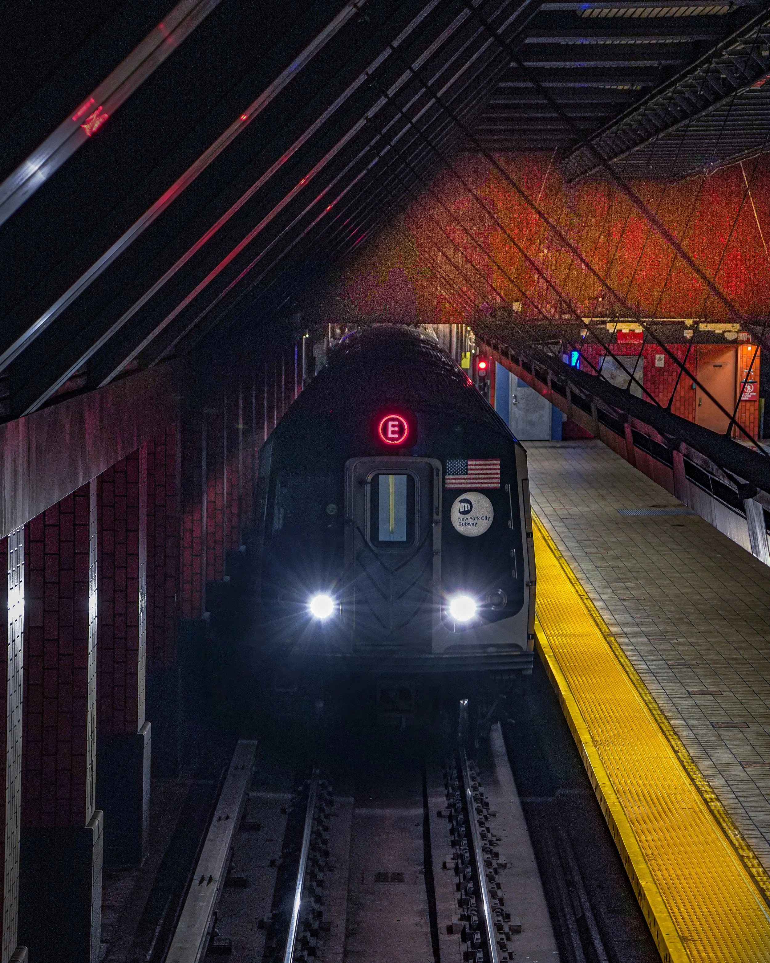A New York City subway train station at night showing a train approaching with its headlights on, the train has an illuminated red letter 'E' on the front and an American flag decal.