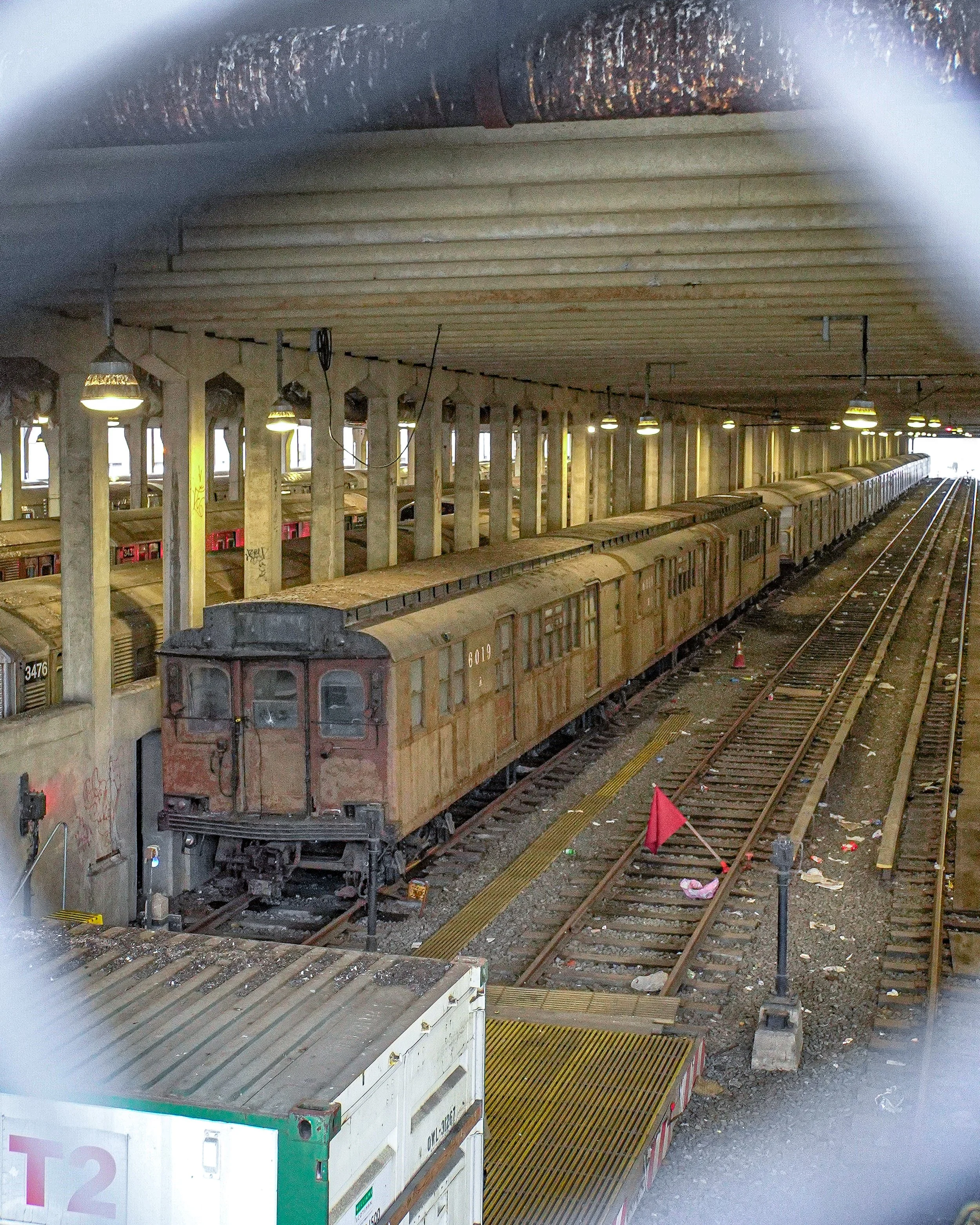 An old, rusty train car in a subway station with graffiti on the wall beside it. The station has a concrete roof with multiple lights hanging from it and yellow safety lines along the platform edges. There is trash along the tracks and a red flag on