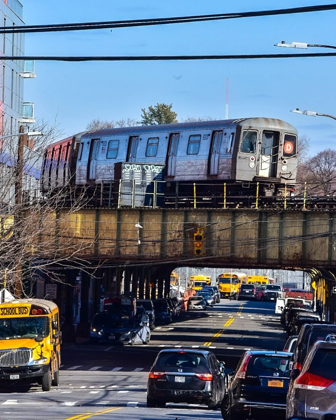 A subway train traveling on an elevated track over a busy city street with parked cars and yellow school buses underneath, and a clear blue sky above.