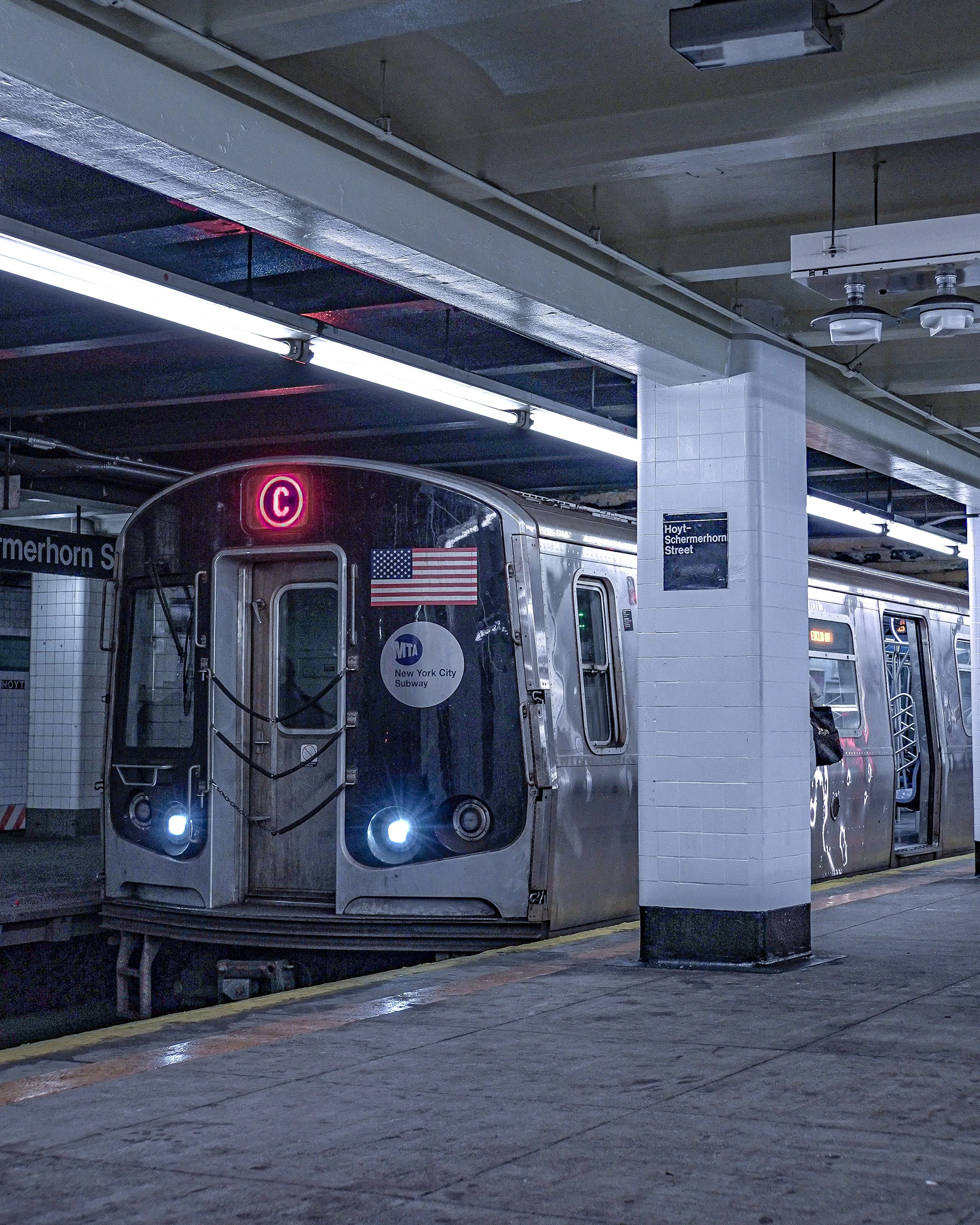A New York City subway train at the Hoyt-Schermerhorn station, with its front illuminated lights and a digital sign showing it is arriving on the C line. The station's sign is visible on the white tiled wall.