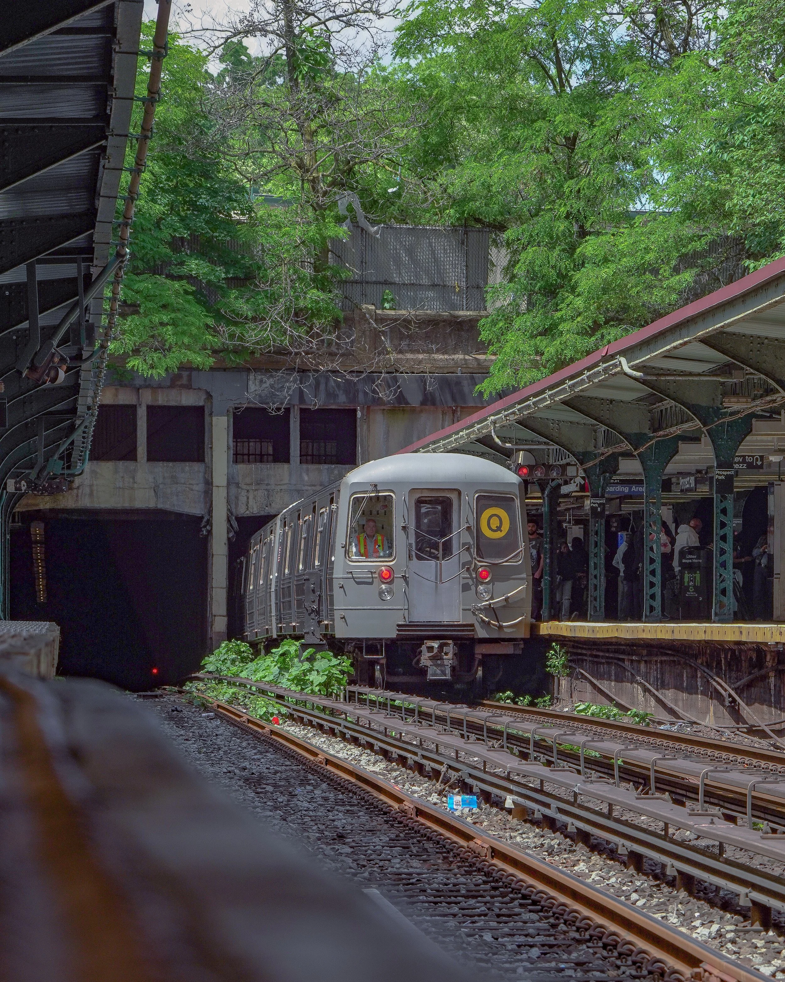 A subway train traveling on an elevated track at a city station during daytime, with people waiting on the platform and green trees in the background.