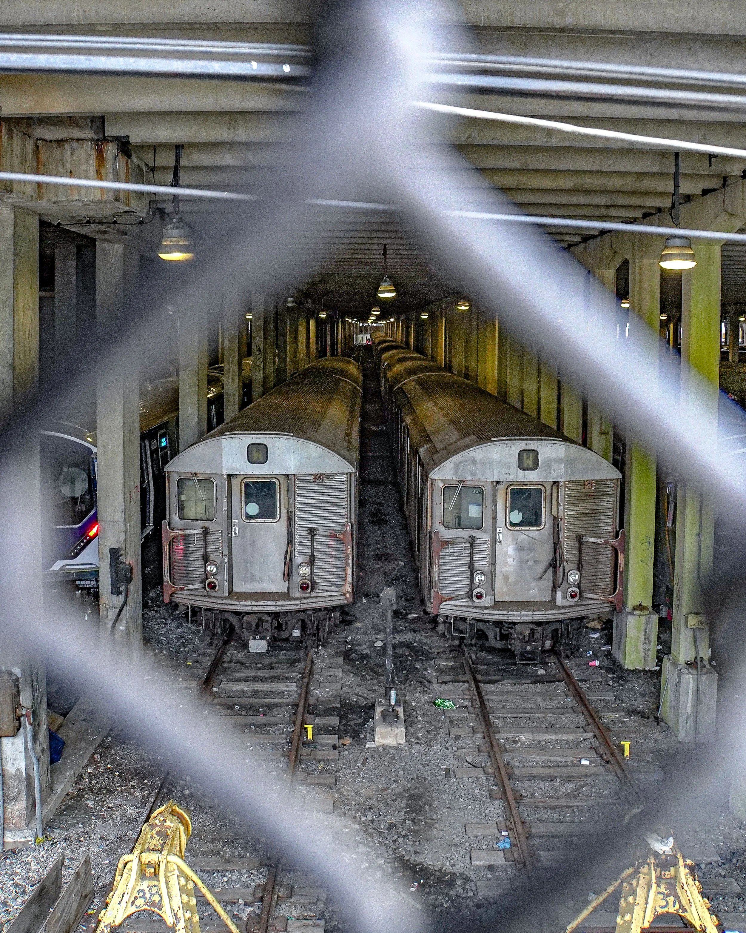 Two old train cars parked on tracks in an underground train yard, viewed through a chain-link fence.