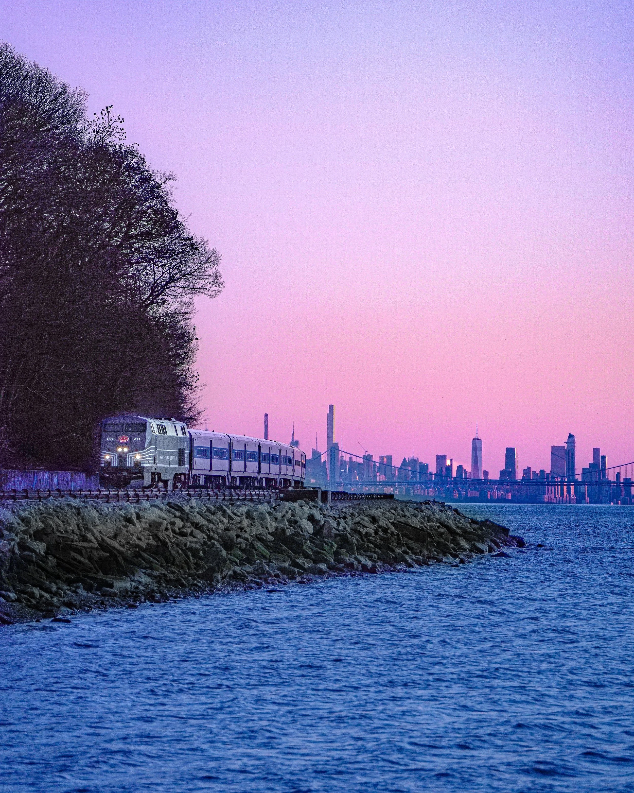 A train traveling along a rocky shoreline with a city skyline and the Brooklyn Bridge in the distance during sunset, with shades of purple and pink in the sky.