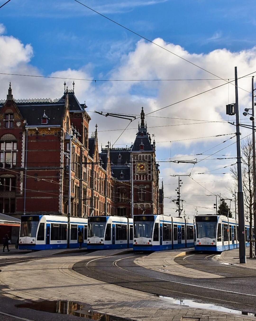 Three modern trams on tracks in front of a historic brick building with a clock tower, power lines overhead, and a partly cloudy sky.