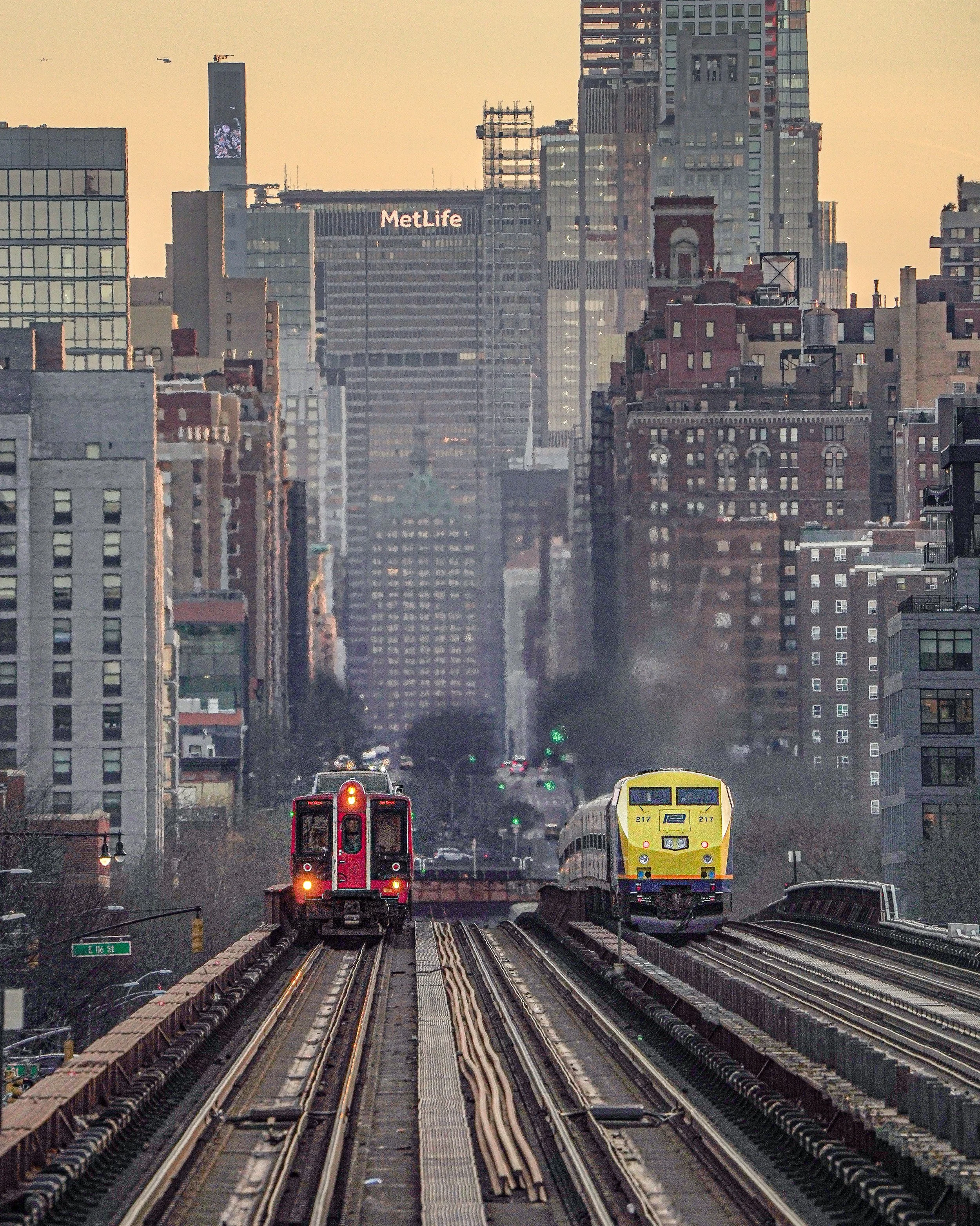 Two trains on elevated tracks in New York City with tall buildings and skyscrapers in the background, including the MetLife building, during sunset.