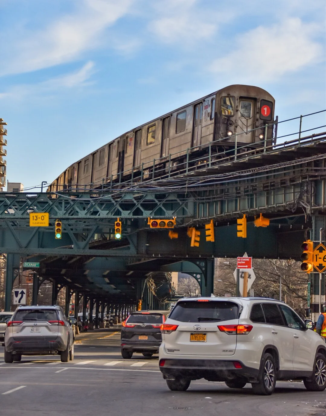 An elevated train traveling on an urban rail line above city streets, with cars and traffic signals below on a clear day.