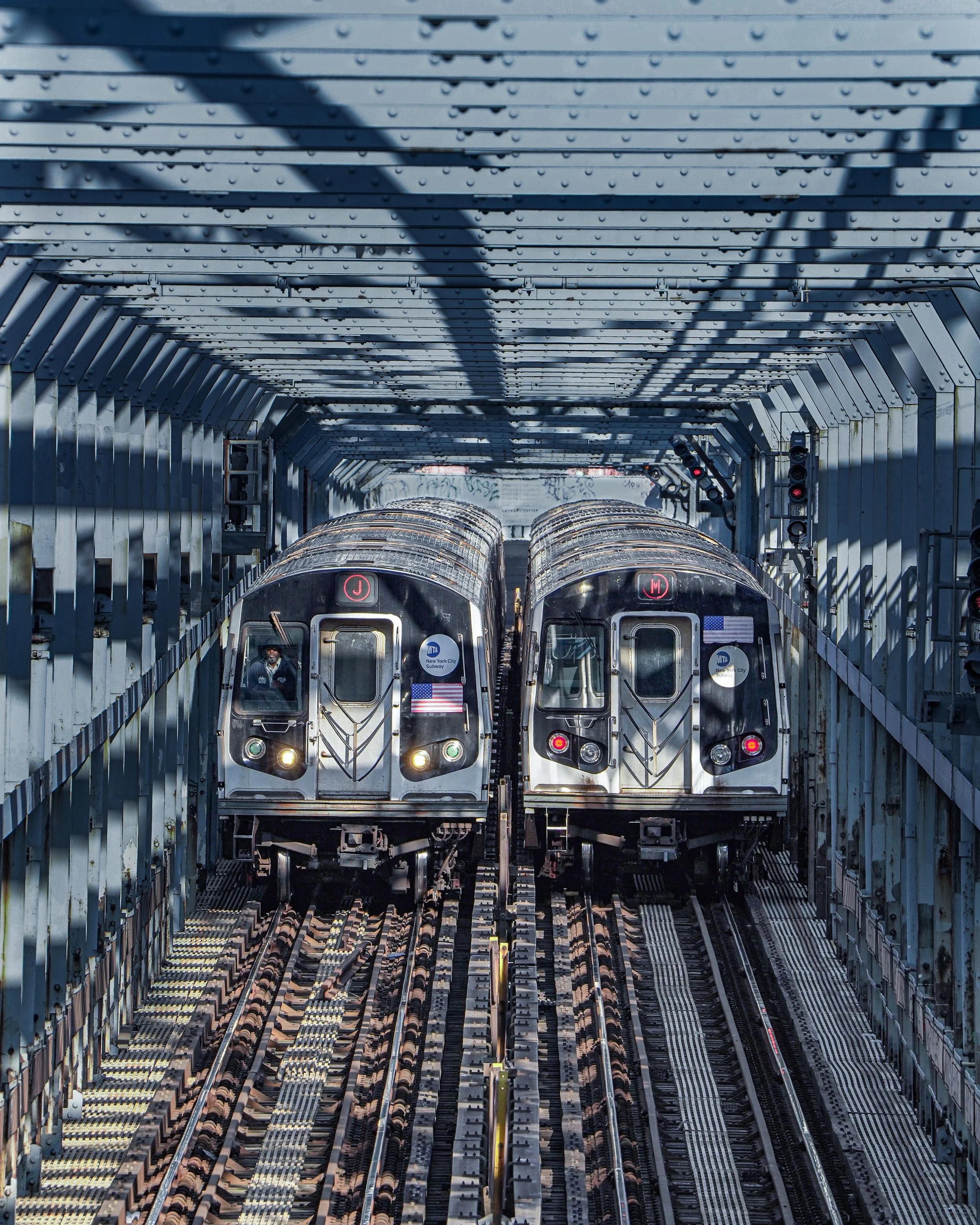 Two New York City subway trains facing each other inside an underground station with metal framework and shadows from the overhead structure.