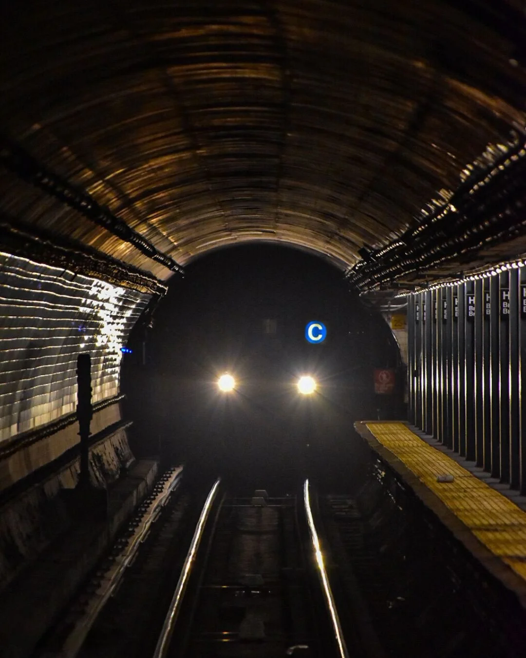 View inside a subway tunnel with a train approaching, showing illuminated headlamps and a blue 'C' sign in the distance.