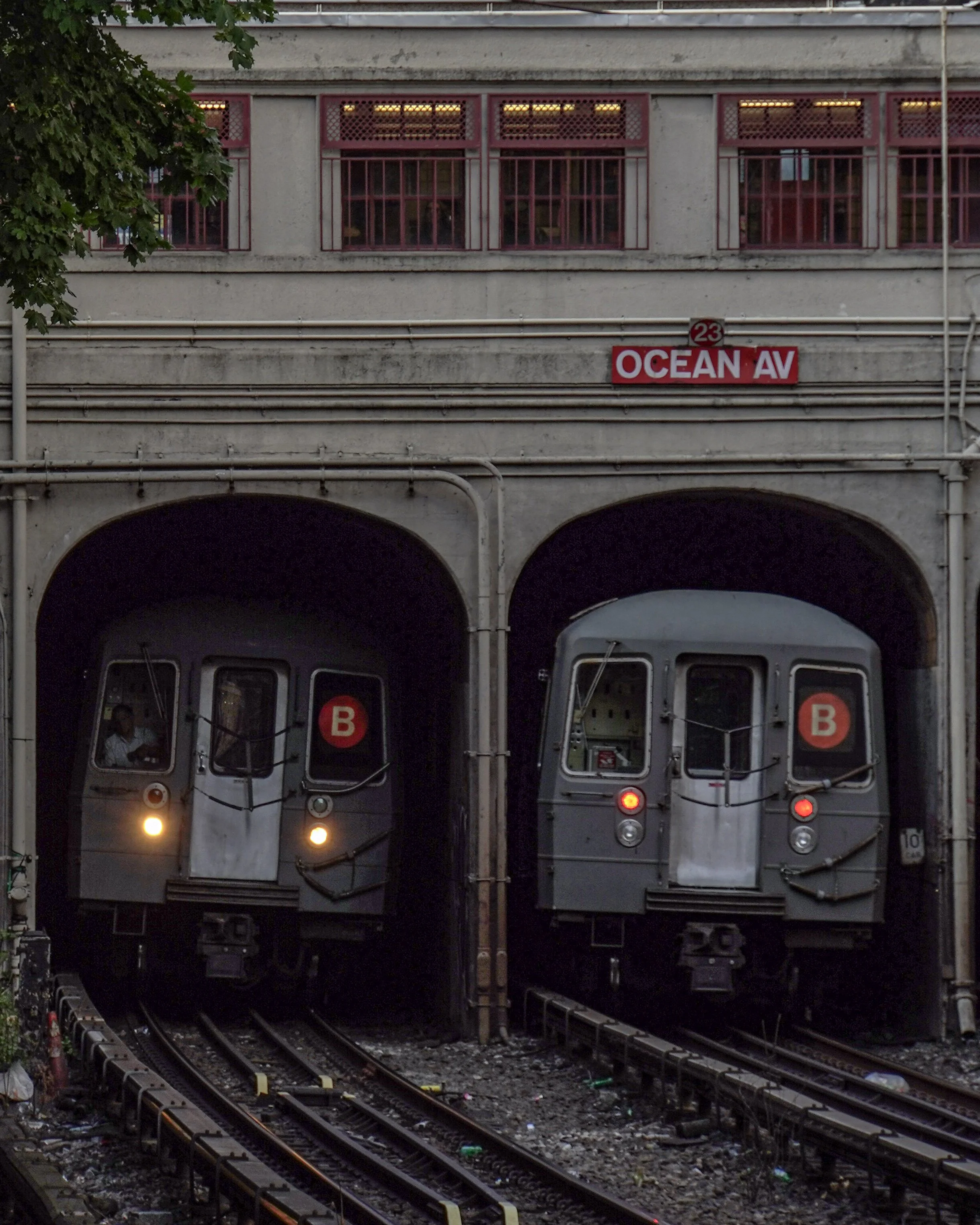 Two subway trains with the B line symbol emerge from tunnels under a concrete building with a pink sign indicating Ocean Avenue.