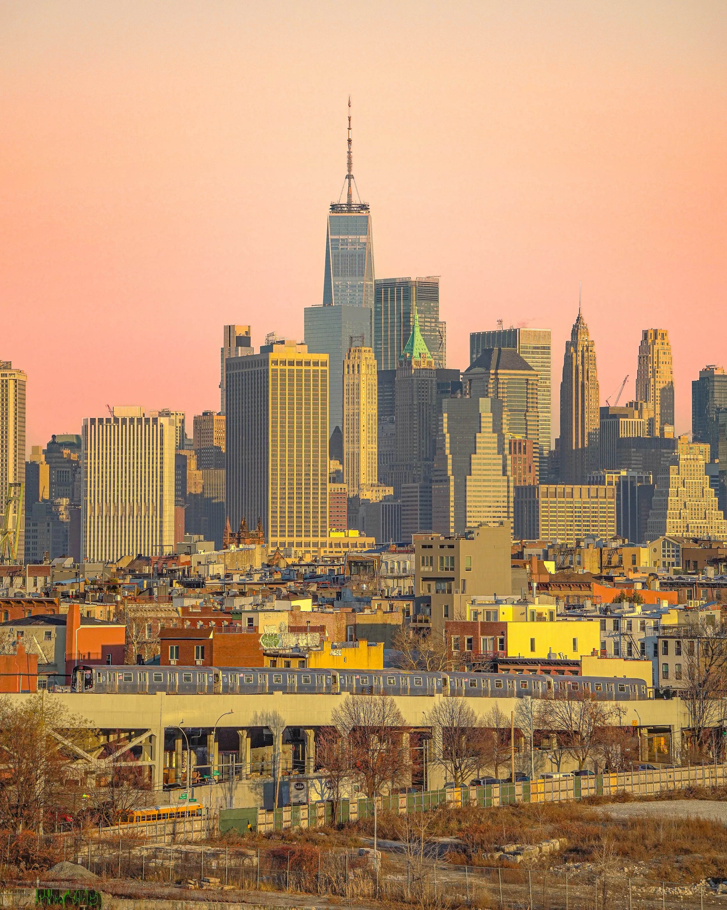City skyline with tall buildings, train on an elevated track in the foreground, and a pinkish sky at sunset.