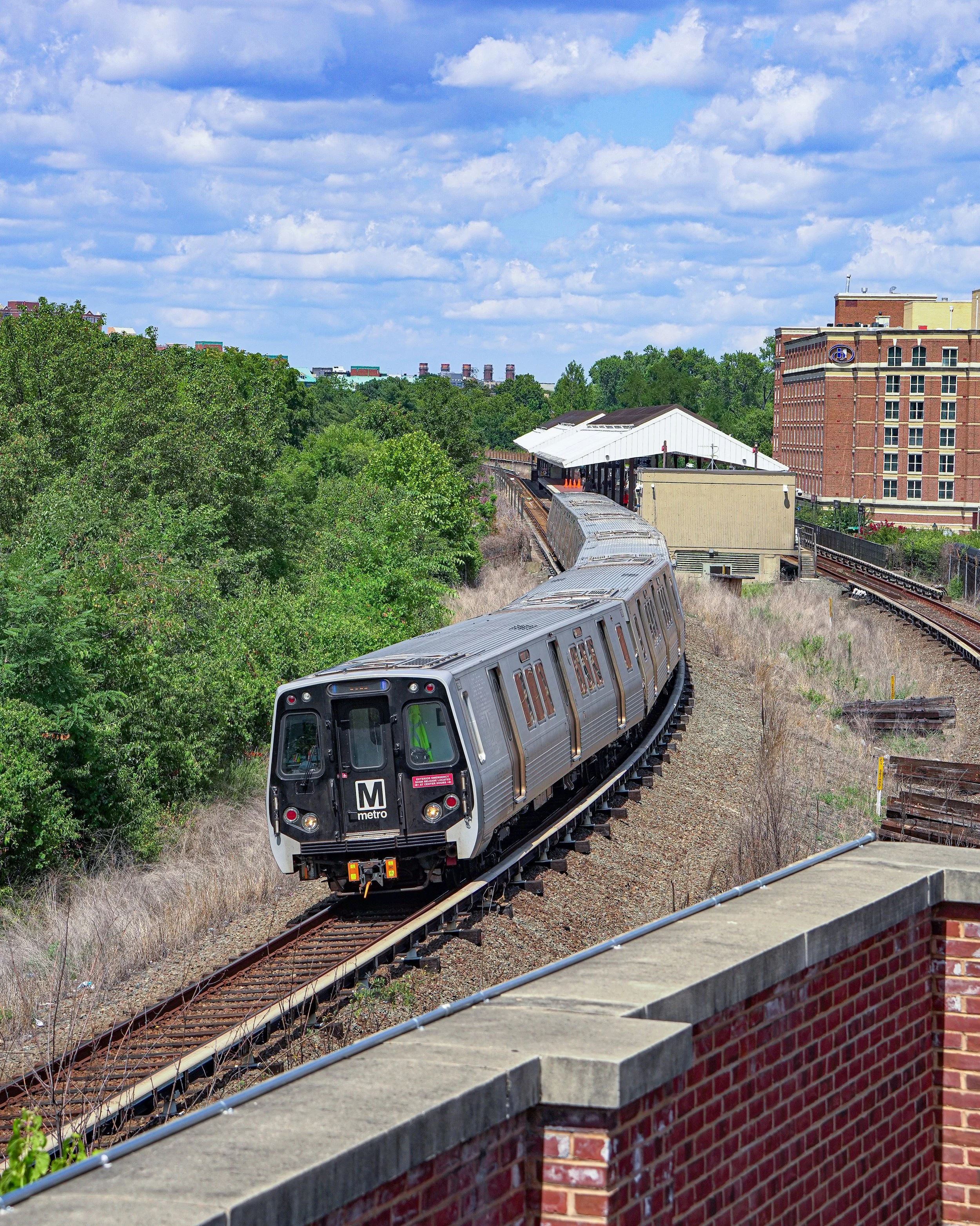 A silver metro train traveling on a curved railway track with green trees on the left, a small white station shelter in the middle, and a tall brick building on the right, beneath a partly cloudy blue sky.