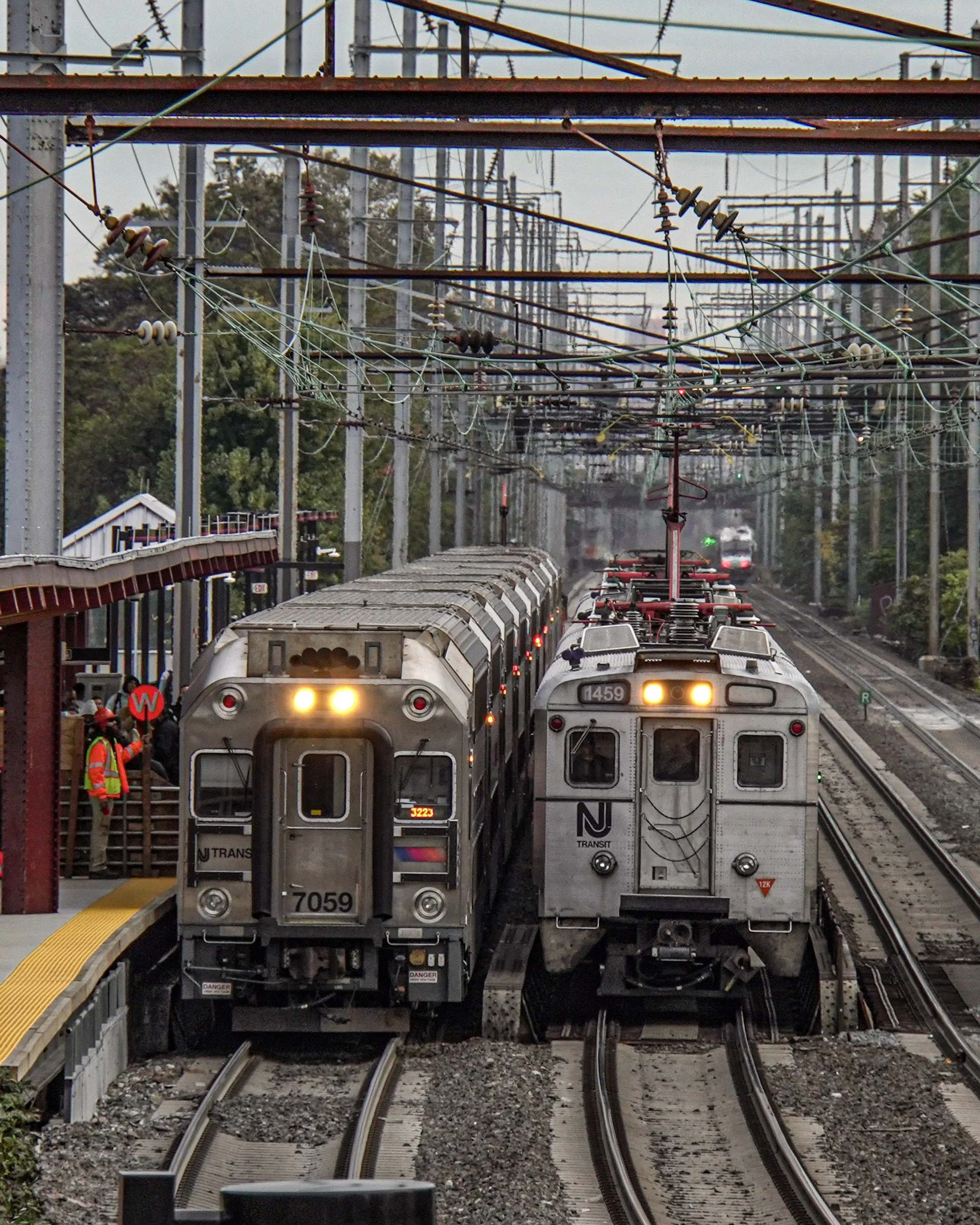 Two subway trains at a station, one with the number 7059 and the other 1459, waiting on separate tracks, with passengers and staff visible near the platform, overhead electrical wires, and trees in the background.