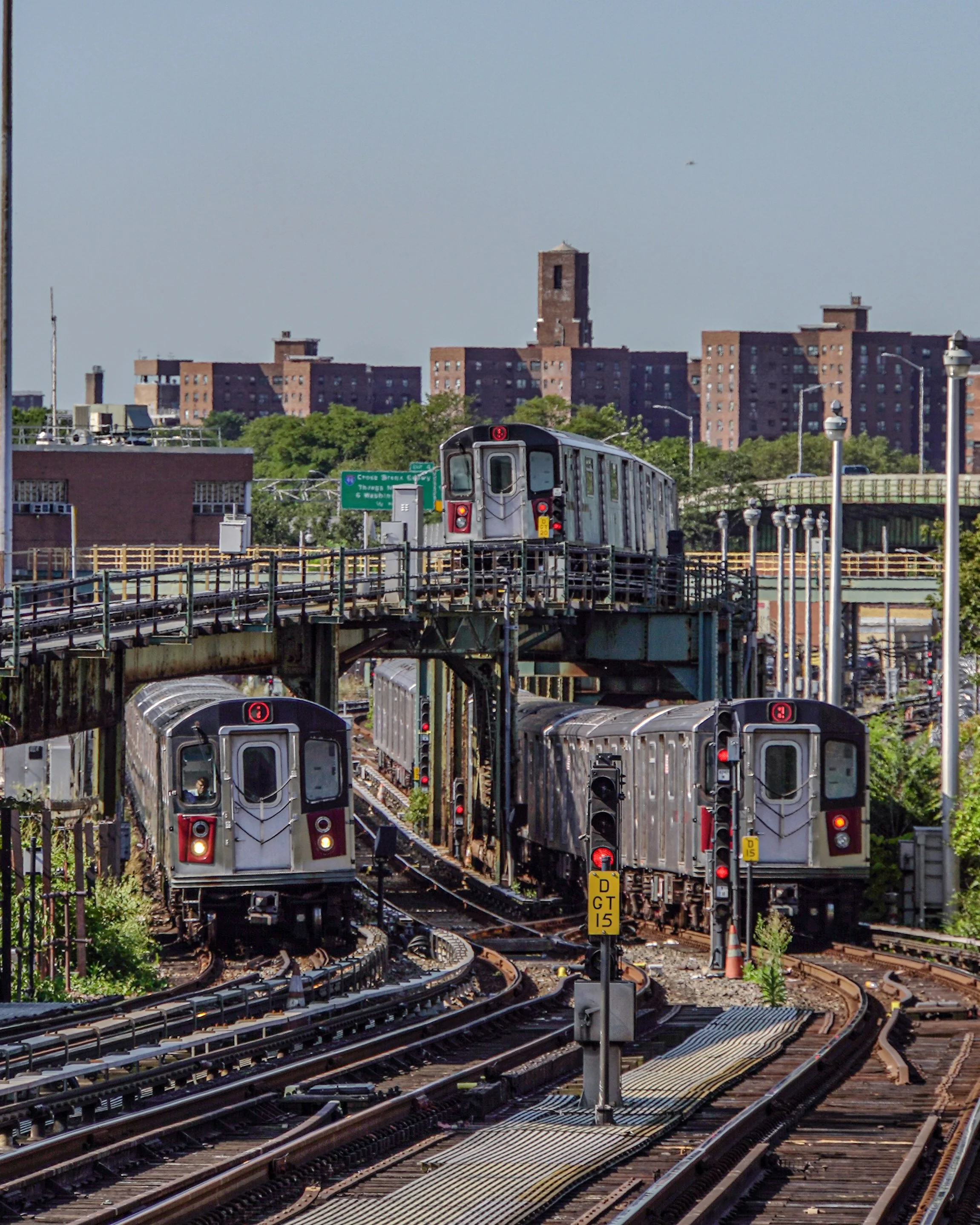 Subway trains on elevated railway tracks in an urban setting with buildings and a bridge in the background.