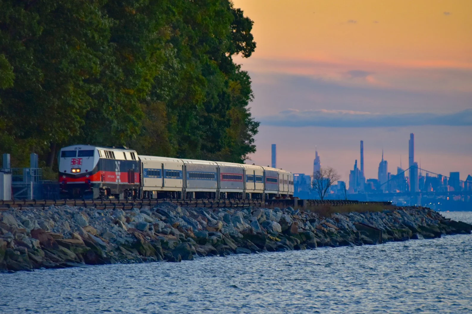 A train traveling along a rocky waterfront with a city skyline and a bridge in the distance at sunset.