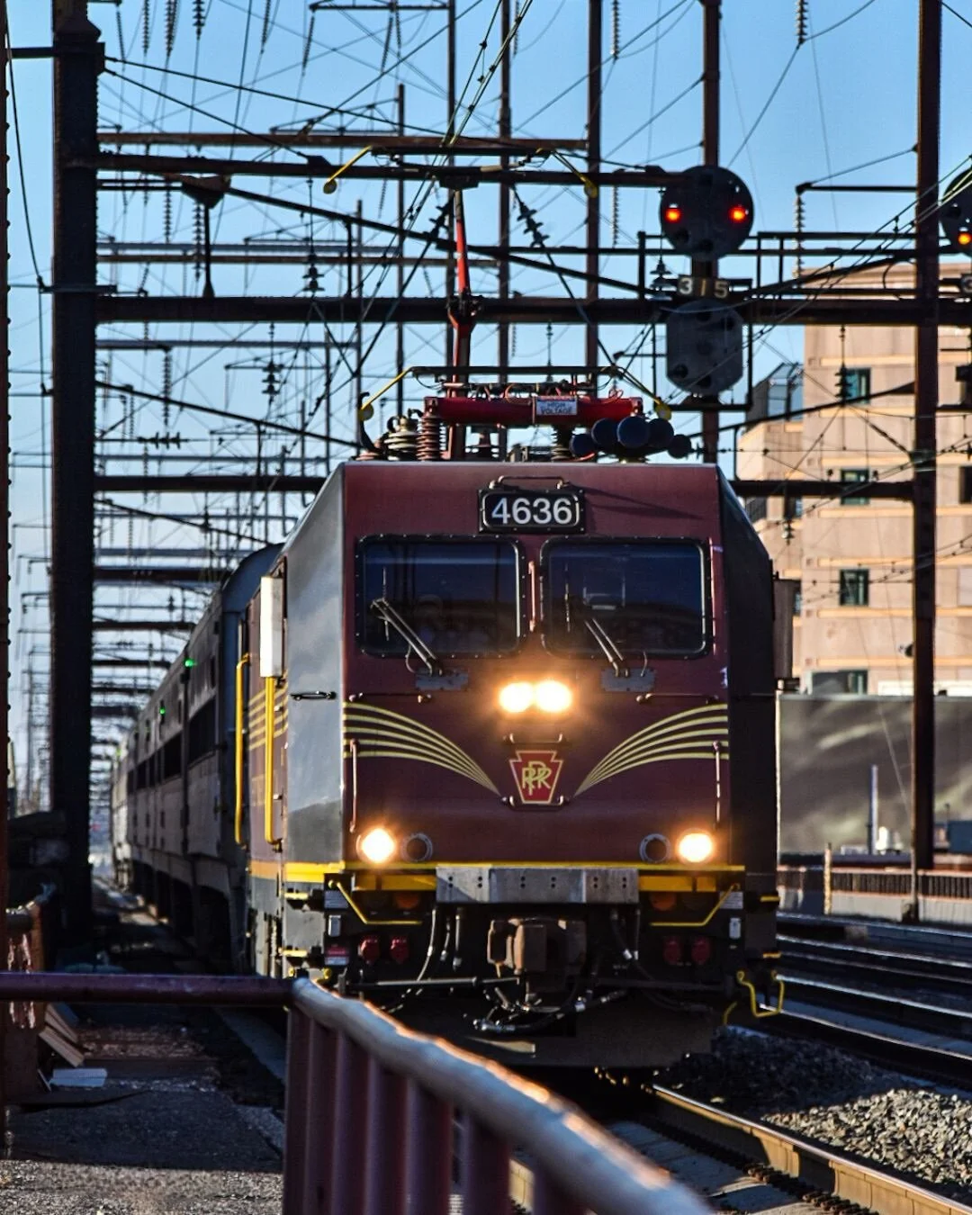 Front view of a maroon and yellow electric train approaching on railway tracks, with numerous overhead power lines and signals overhead, and a building in the background.