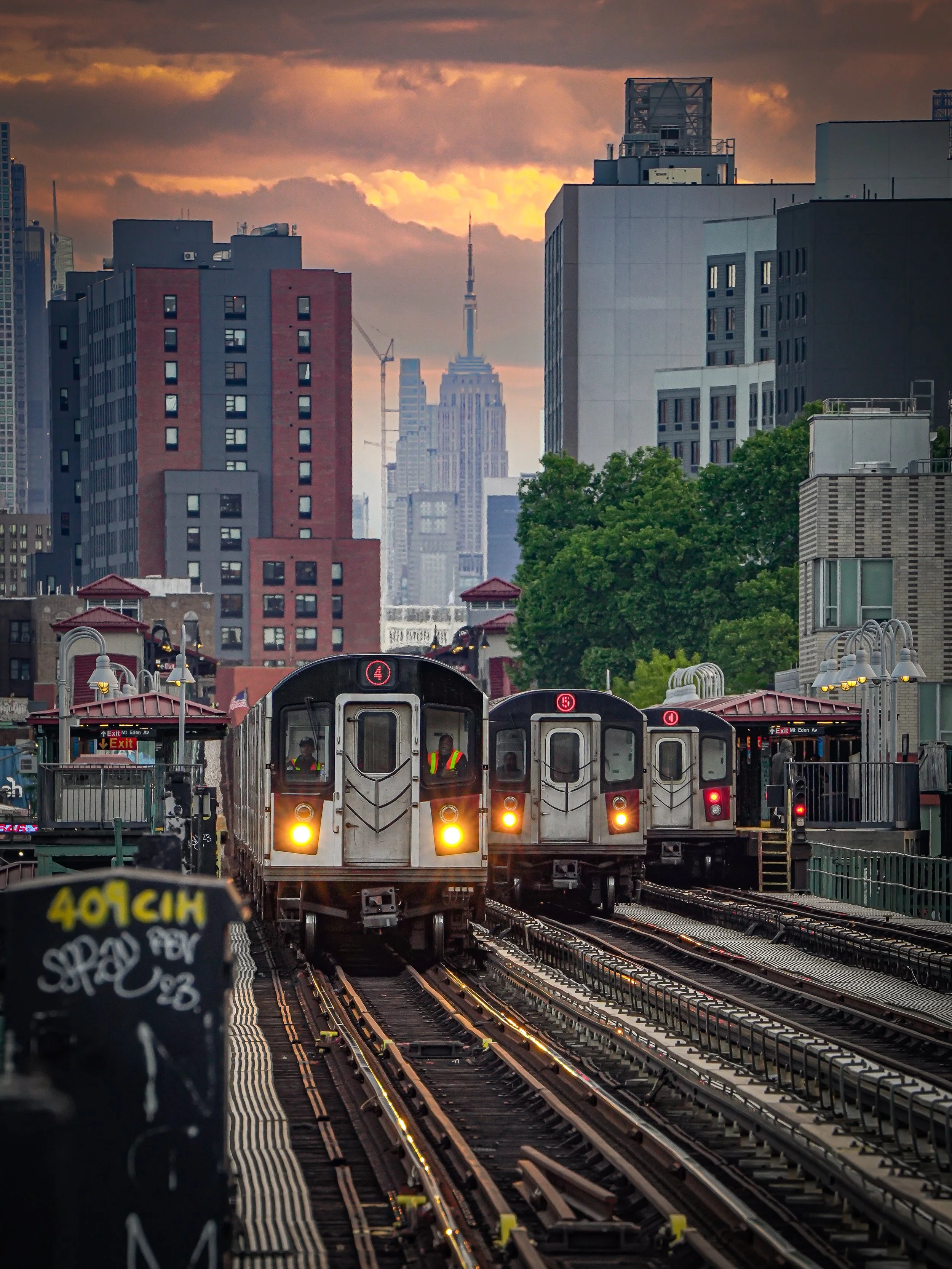Subway trains on elevated tracks in New York City with tall buildings and the Empire State Building visible in the background during sunset.