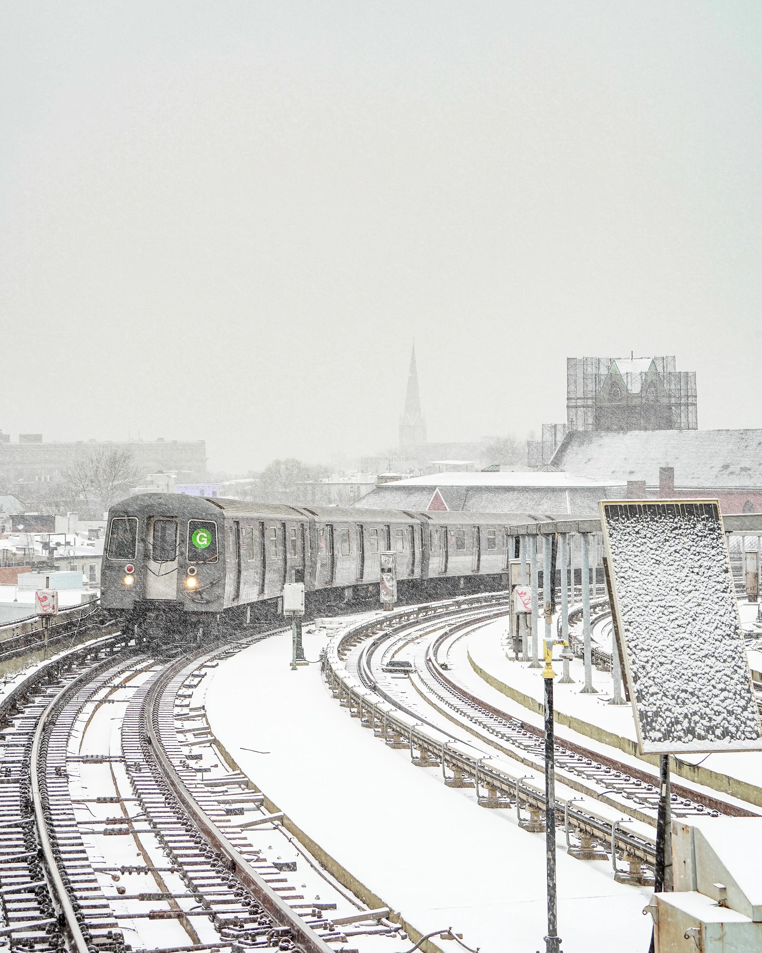 A train moving on snow-covered tracks in a city, with a sign covered in snow and a faint skyline in the background.