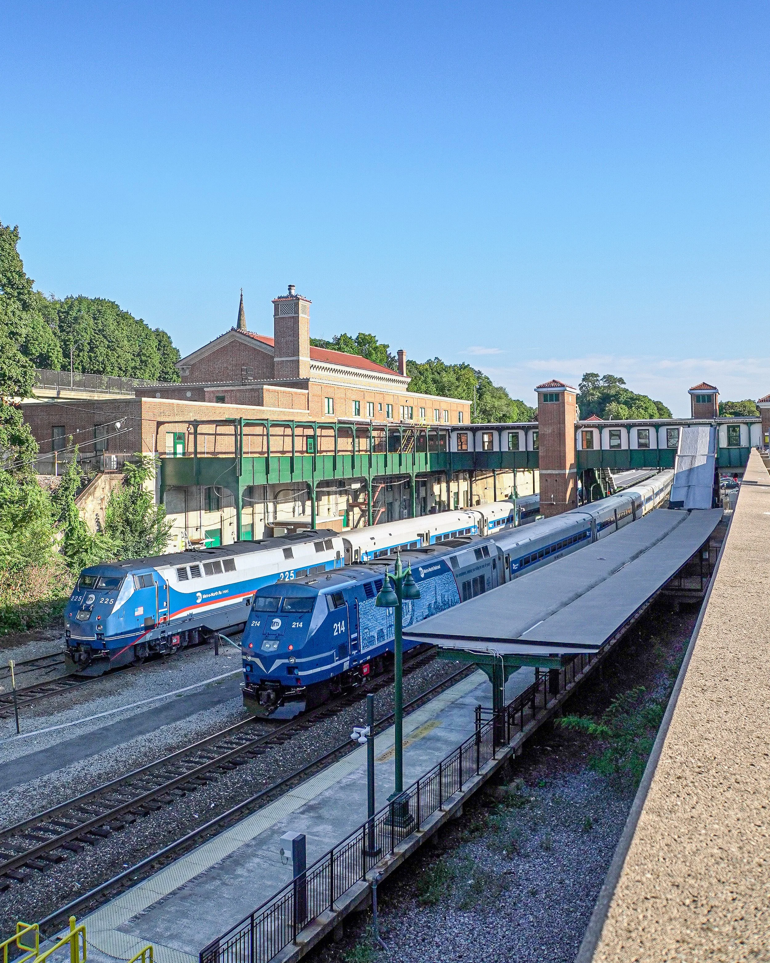Two Amtrak trains parked at a station with a historic brick building and green trees in the background under a clear blue sky.