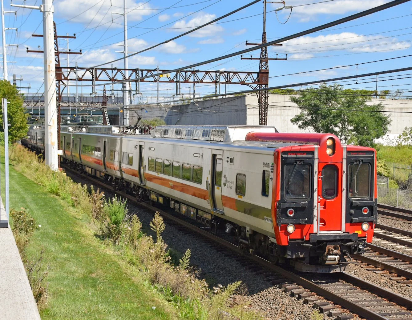 A modern commuter train with a red front and white body with orange and gray stripe runs on railroad tracks underneath overhead electric wires on a clear, sunny day.