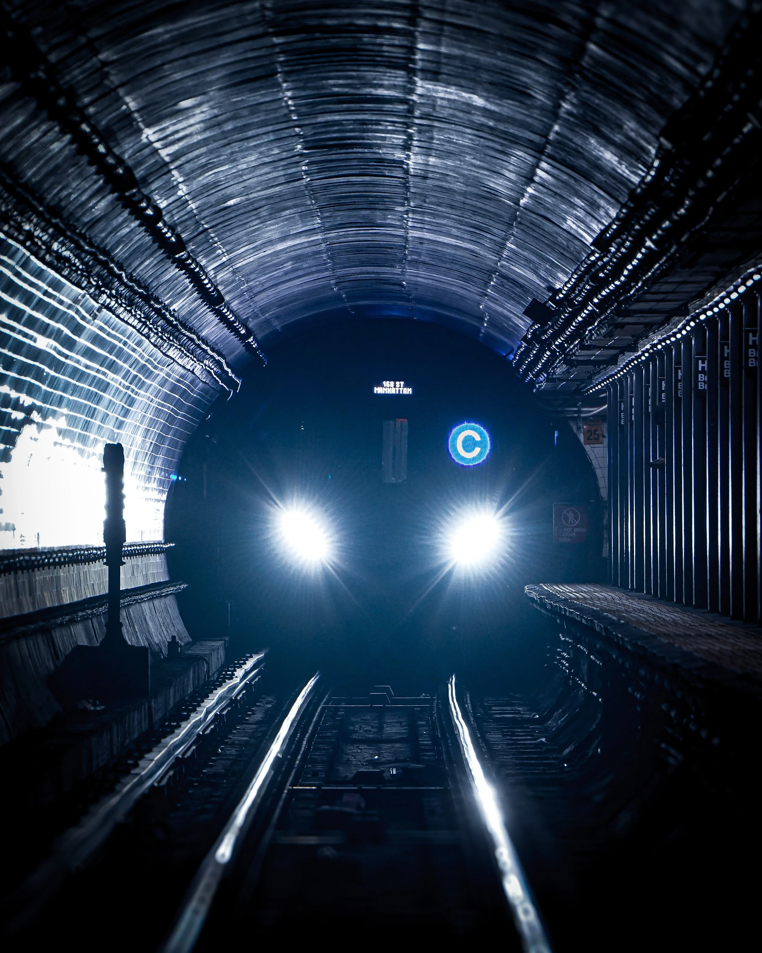 A train approaching through a dark underground tunnel with bright headlights, illuminated blue and white signs, and glowing track lines.