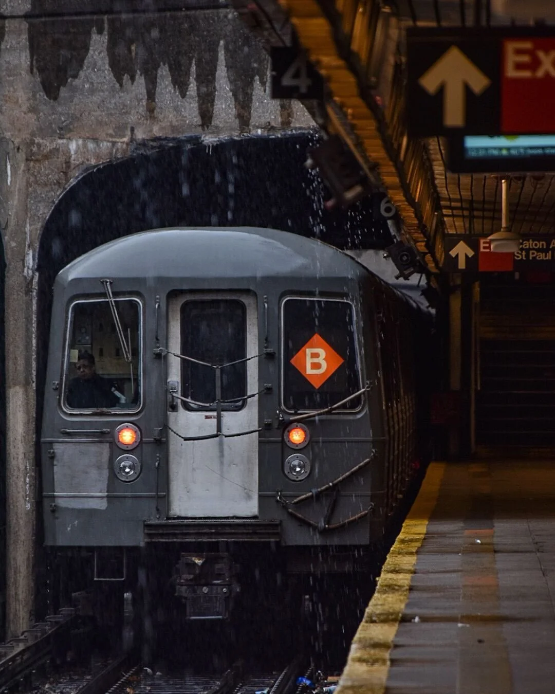 A subway train emerging from a tunnel at a dimly lit station, with rain-like water droplets falling and visible underground infrastructure.